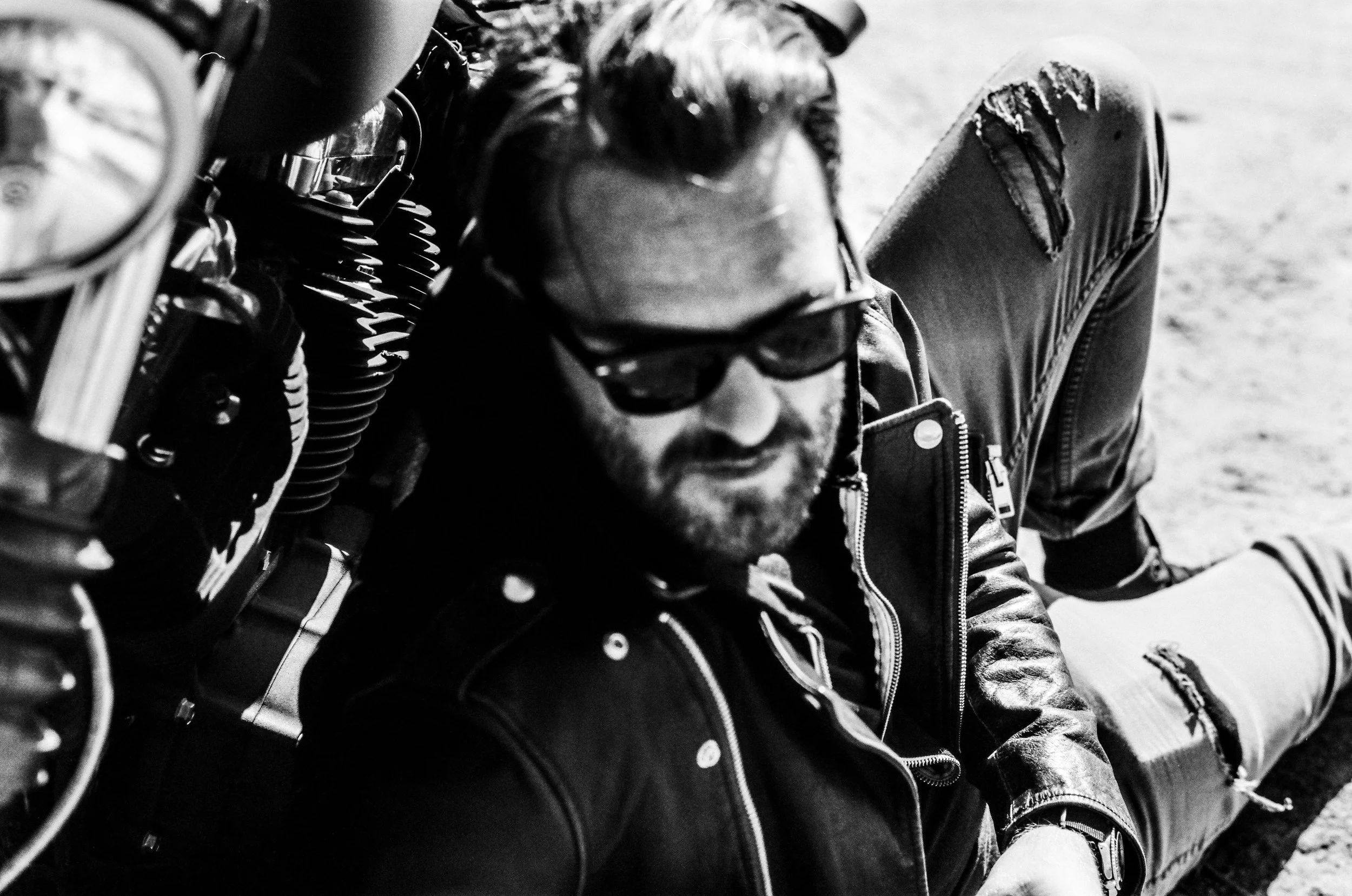 Black and white photo of three people sitting close together outdoors, wearing sunglasses and ripped jeans, possibly at the beach or in a similar setting.