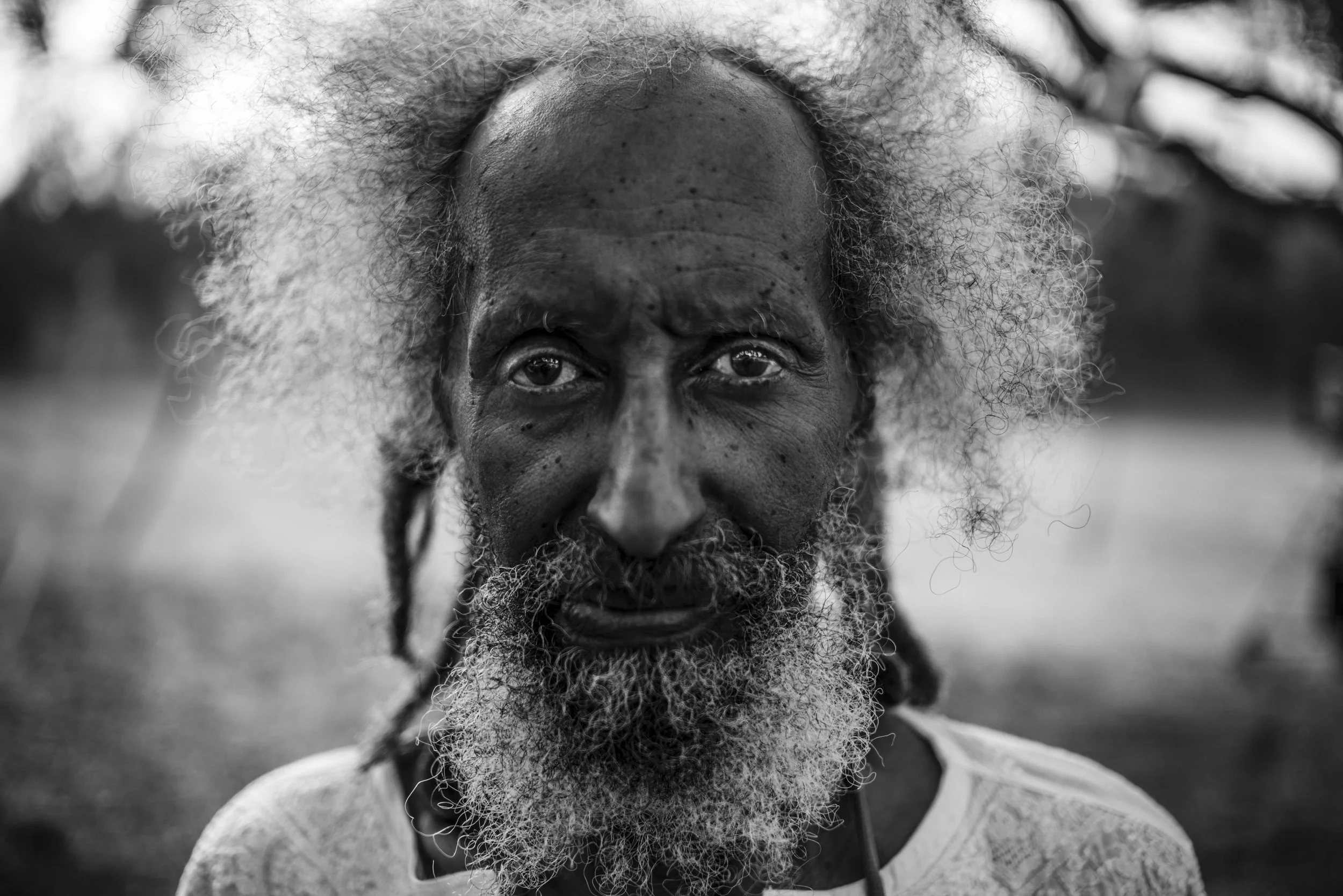 Close-up black and white photo of an elderly man with a full white beard and curly hair, smiling outdoors.