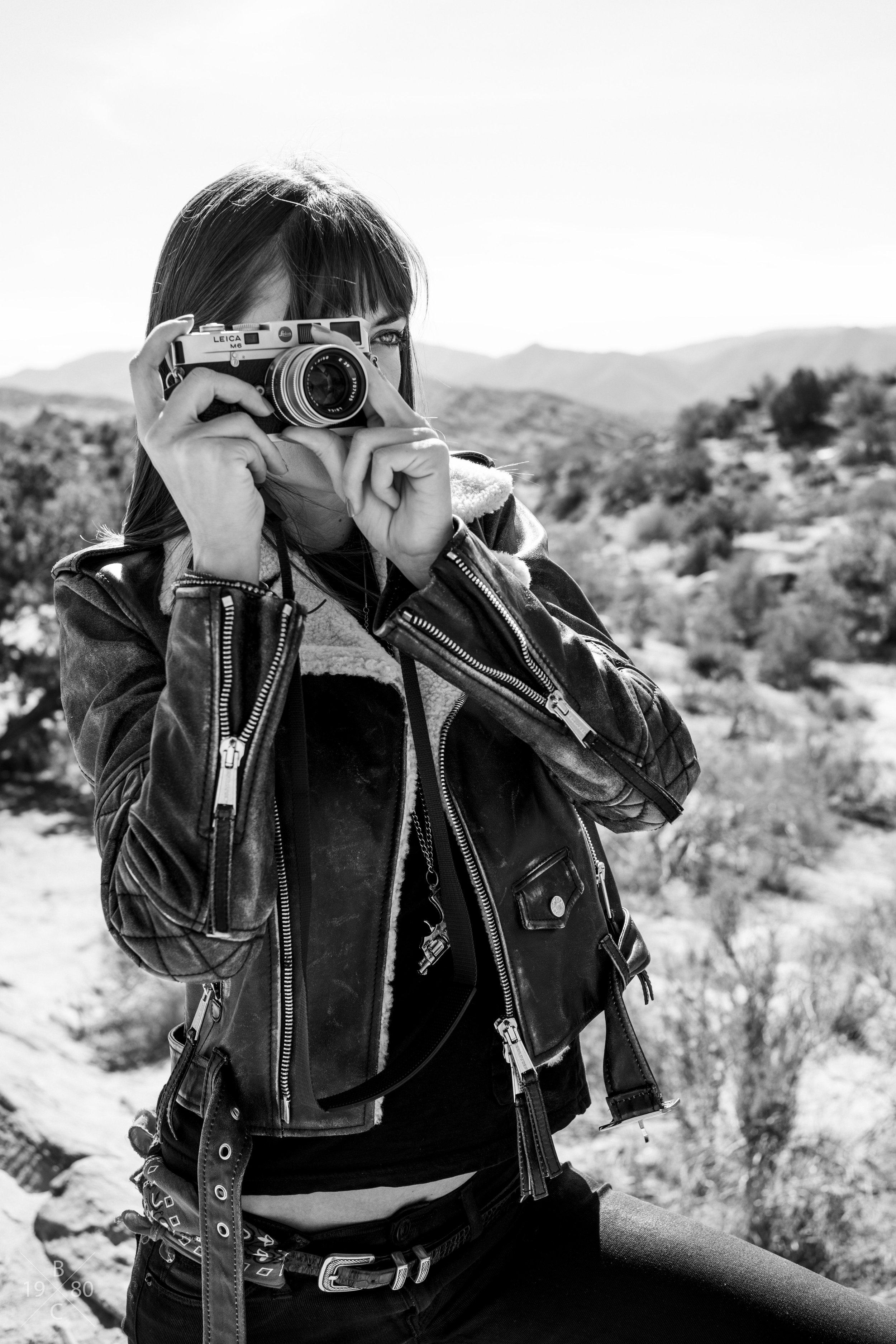 A woman with dark hair in a leather jacket taking a photograph with a vintage Leica camera outdoors in a desert landscape with mountains in the background.