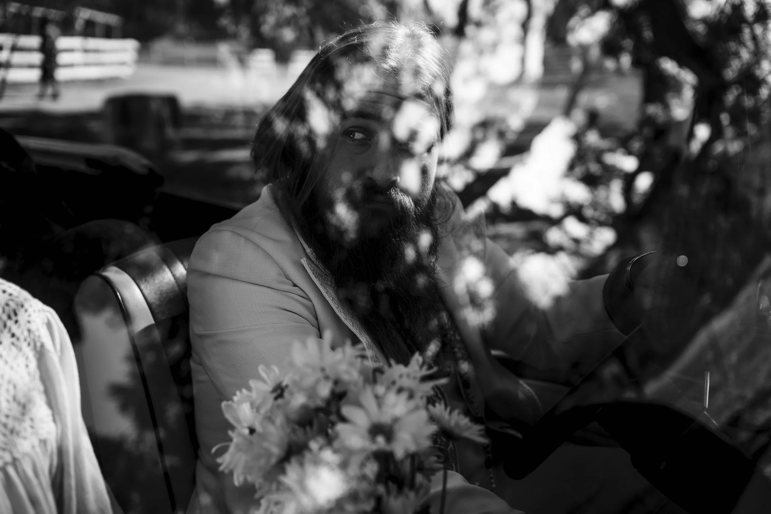 Black and white photo of a man with long hair and a beard, partially obscured by reflections or shadows, sitting outdoors with trees and a bench in the background.
