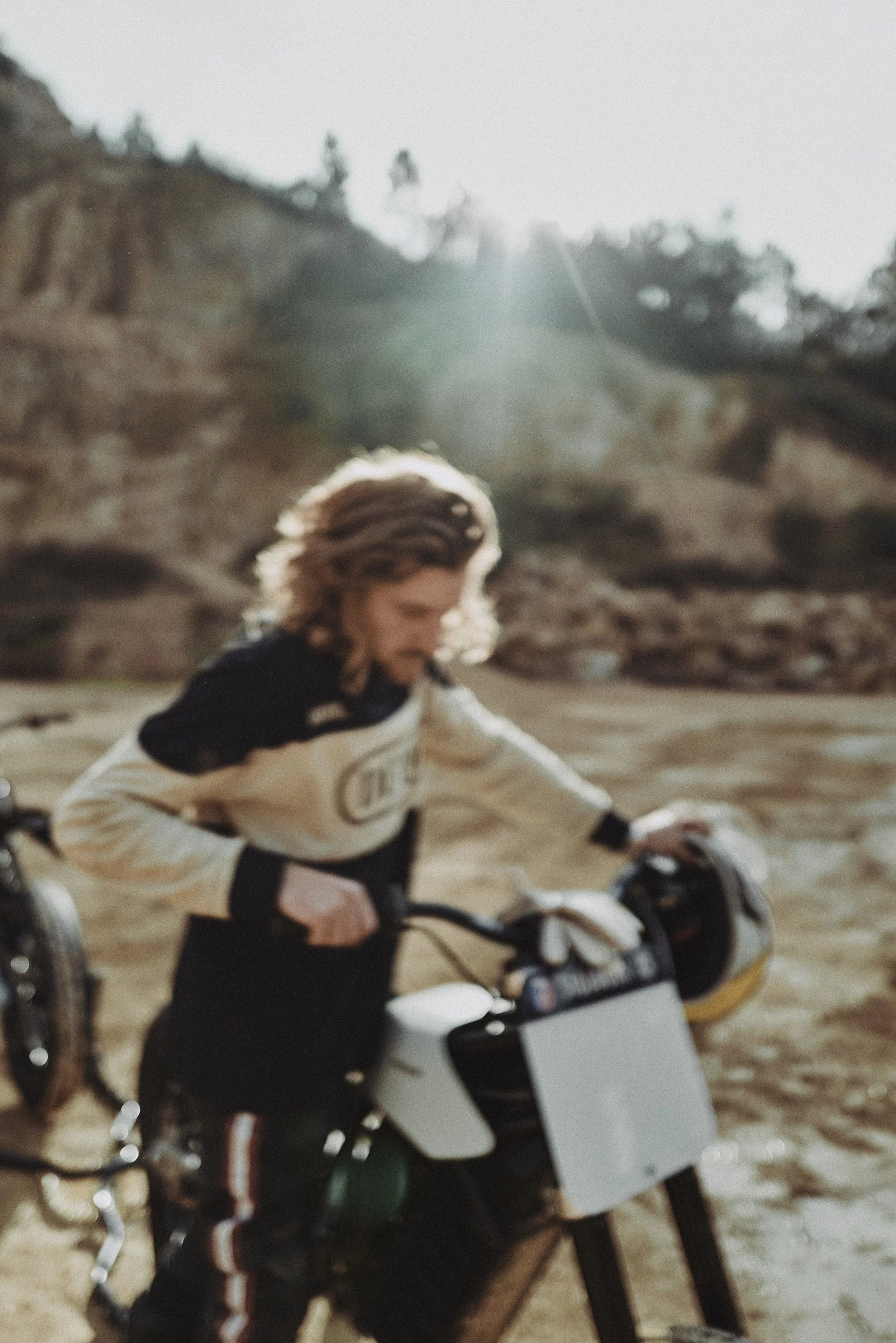 A person riding a motorcycle on a dirt path with a rocky hillside and trees in the background. The person is wearing a beige and black jacket and has wavy hair.