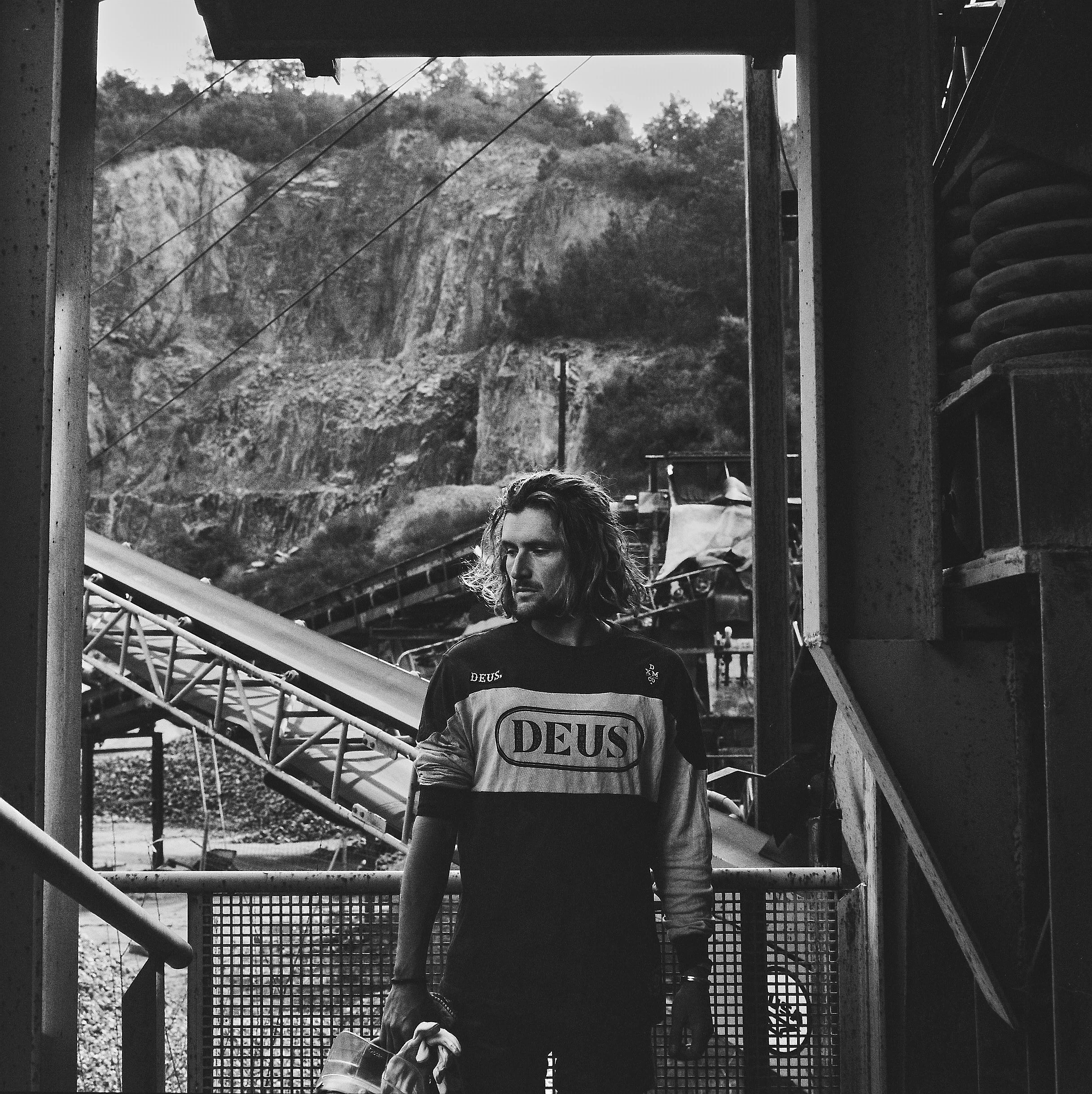 A young man with long hair and a beard standing inside a wooden structure, looking out through a frame with a mountain and trees in the background. He is wearing a shirt with the brand name 'DEUS' on it.