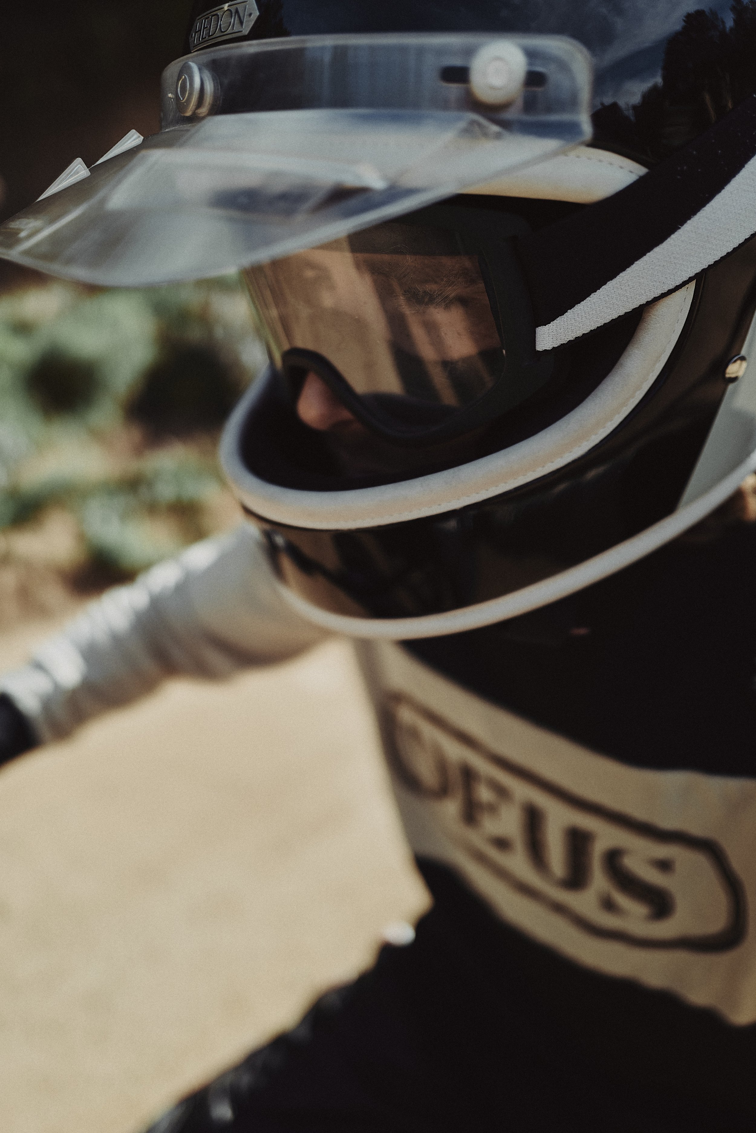 Close-up of a black motorcycle helmet with a clear visor, reflecting sky and trees, and a white stripe on the side, placed on a beige surface.