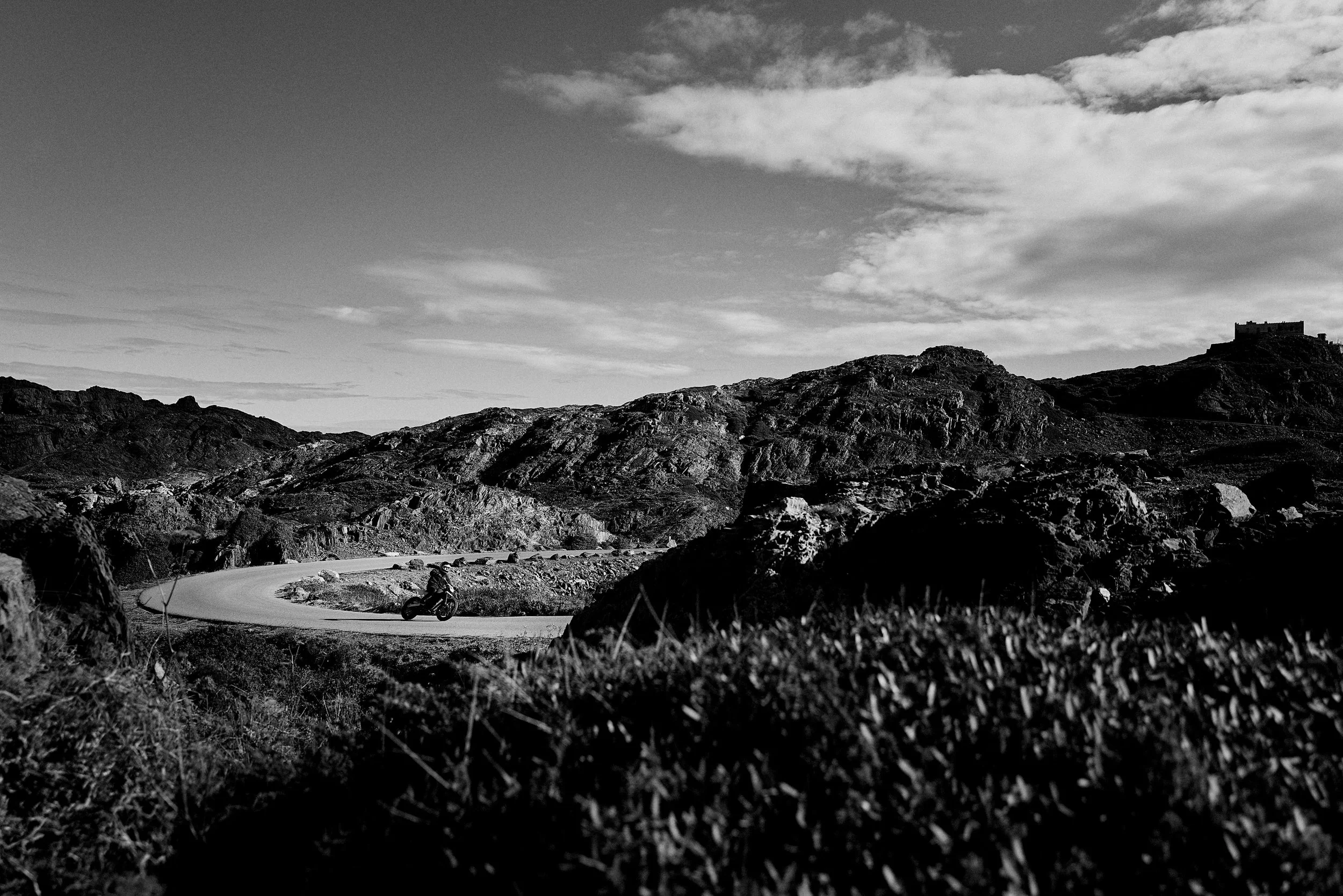 A person riding a motorcycle on a winding mountain road with rugged terrain, hills, and a castle in the distance under a partly cloudy sky.