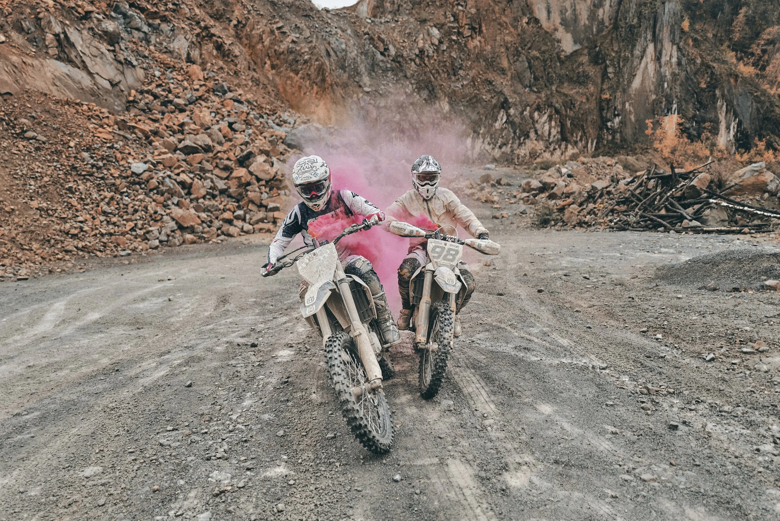 Two dirt bike riders wearing helmets and protective gear riding on a rugged dirt trail surrounded by rocky terrain and high cliffs, with pink smoke emitted from their bikes.