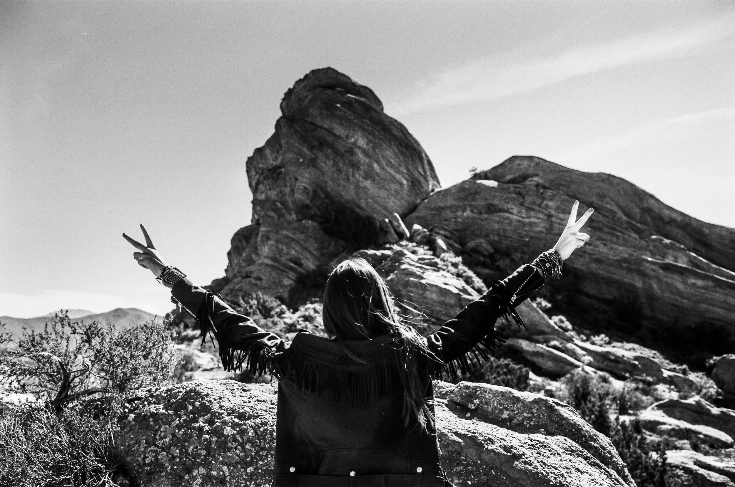 Person in a jacket making a peace sign with both hands in front of rock formations in a desert landscape