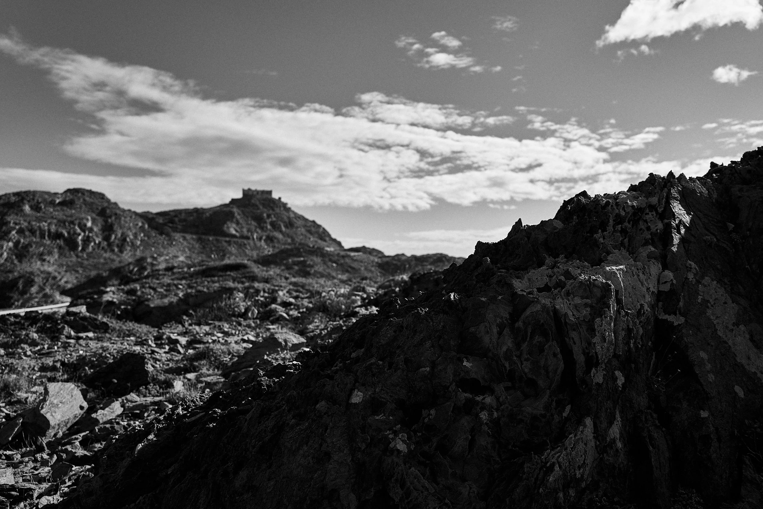 A black and white landscape photo featuring a mountain with a castle or fortress on top, rugged terrain in the foreground, and clouds in the sky.
