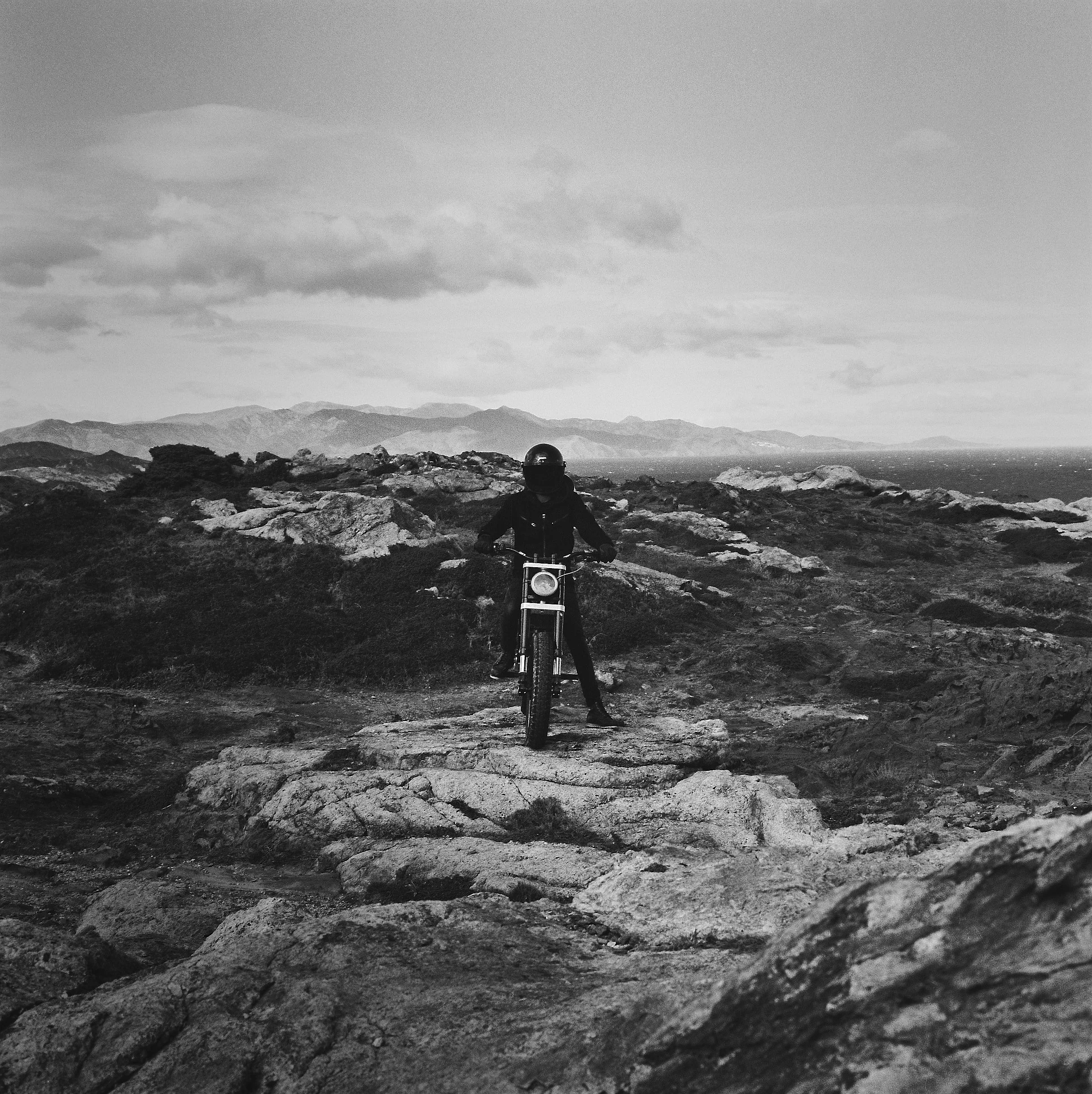 A person riding a motorcycle on a rough, rocky terrain with mountains in the background and a cloudy sky above.