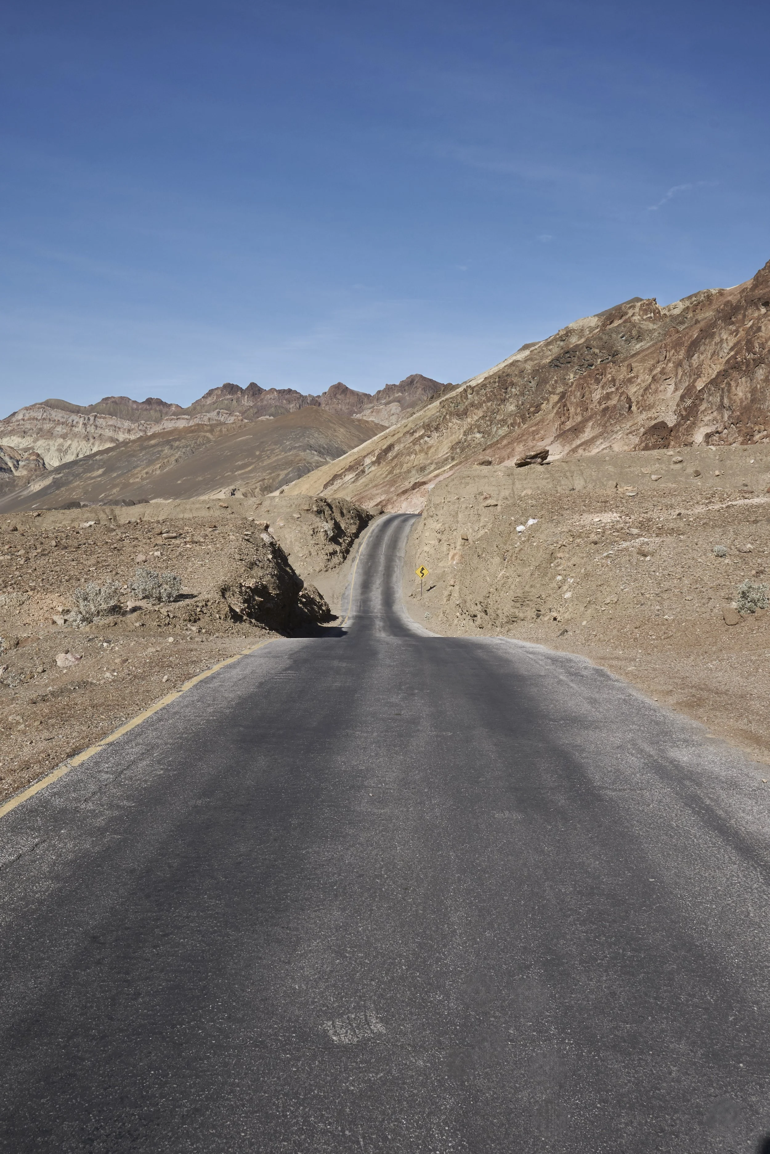 A paved road in a desert landscape with brown mountains and a partly cloudy blue sky.