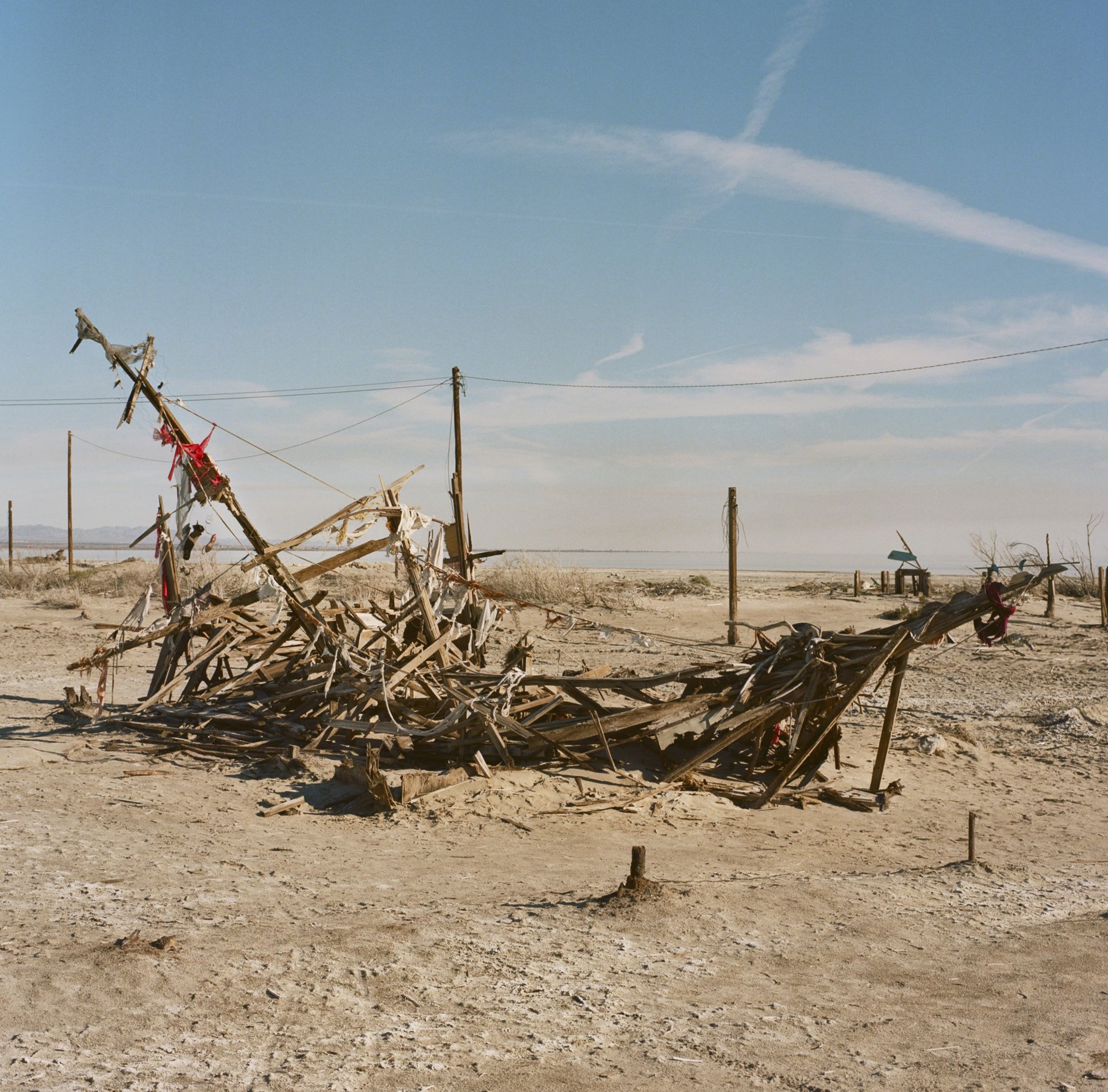 A desert landscape with remnants of a burned wooden structure, power lines in the background, and a clear blue sky with some clouds.
