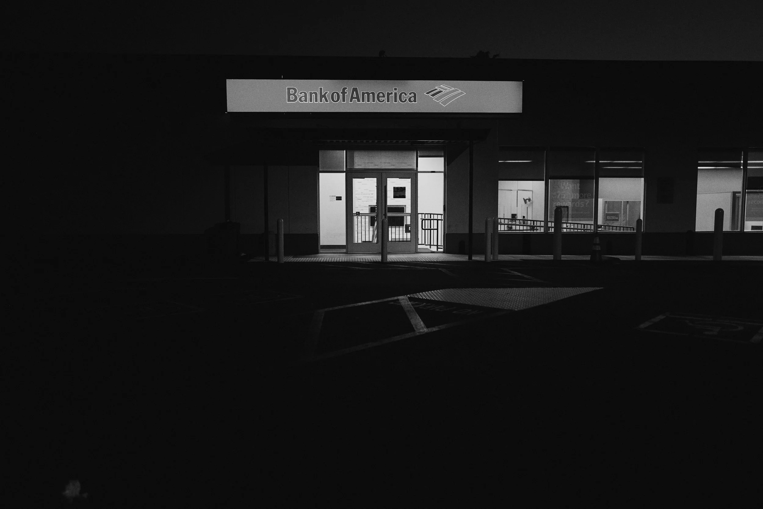 A black and white image of the entrance to a Bank of America branch at night. The bank signage is illuminated above the door, and the building's glass door and windows are visible. The parking lot in front is dark with painted lines and a few bollard