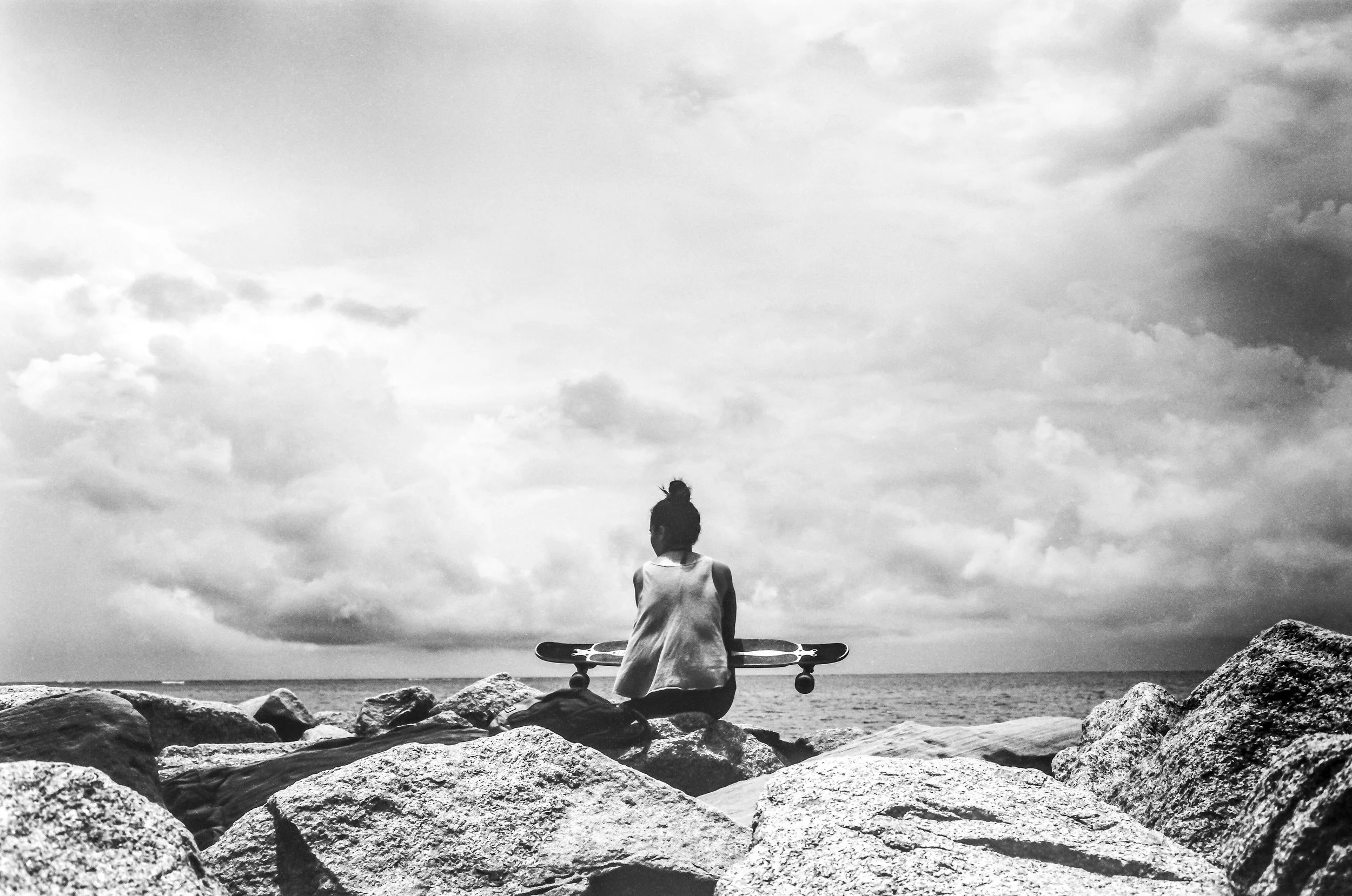 A person sitting on rocks by the water, gazing at the cloudy sky, with a skateboard resting against their knees.