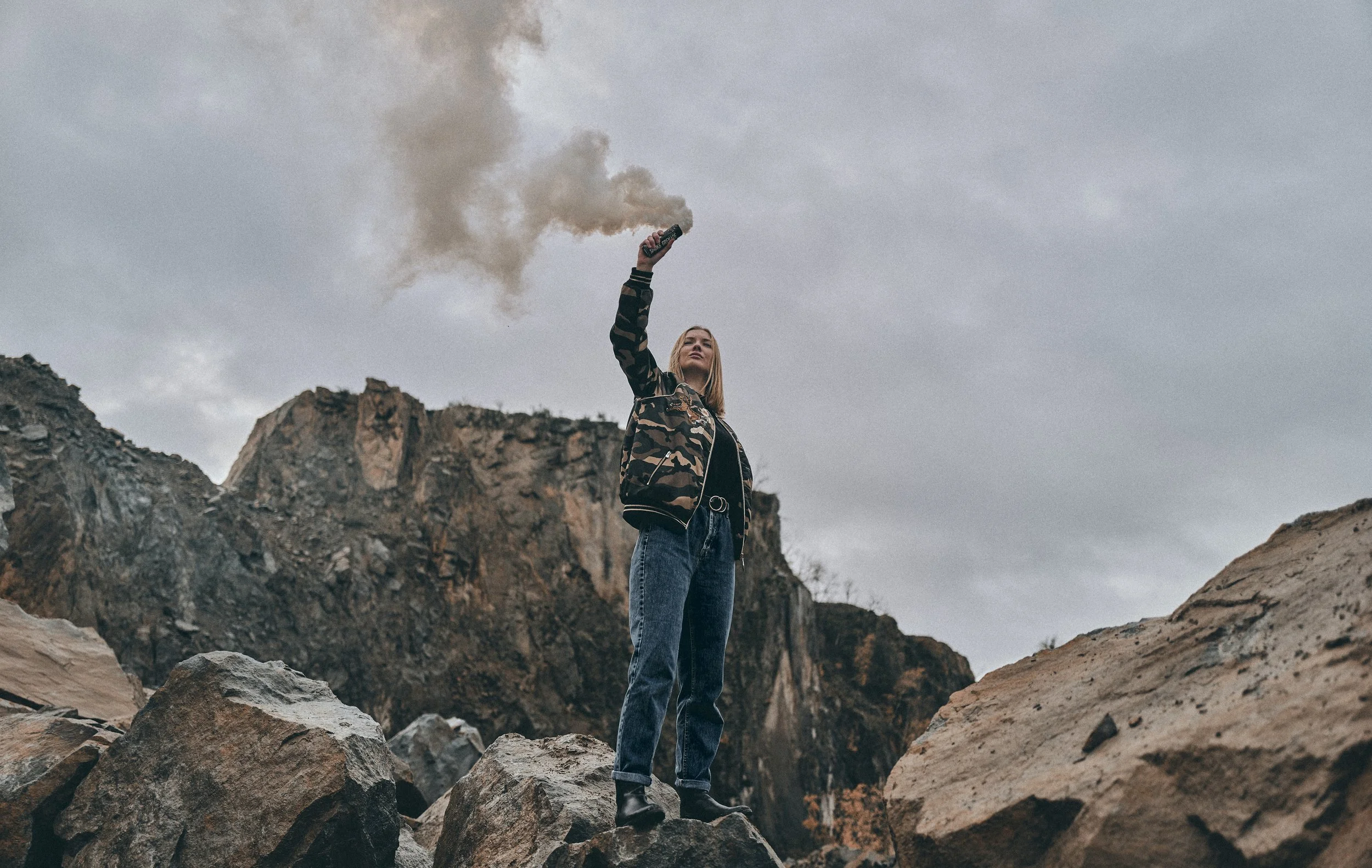 A woman standing on rocky terrain in a mountainous area, holding a smoke flare that emits smoke into the cloudy sky.