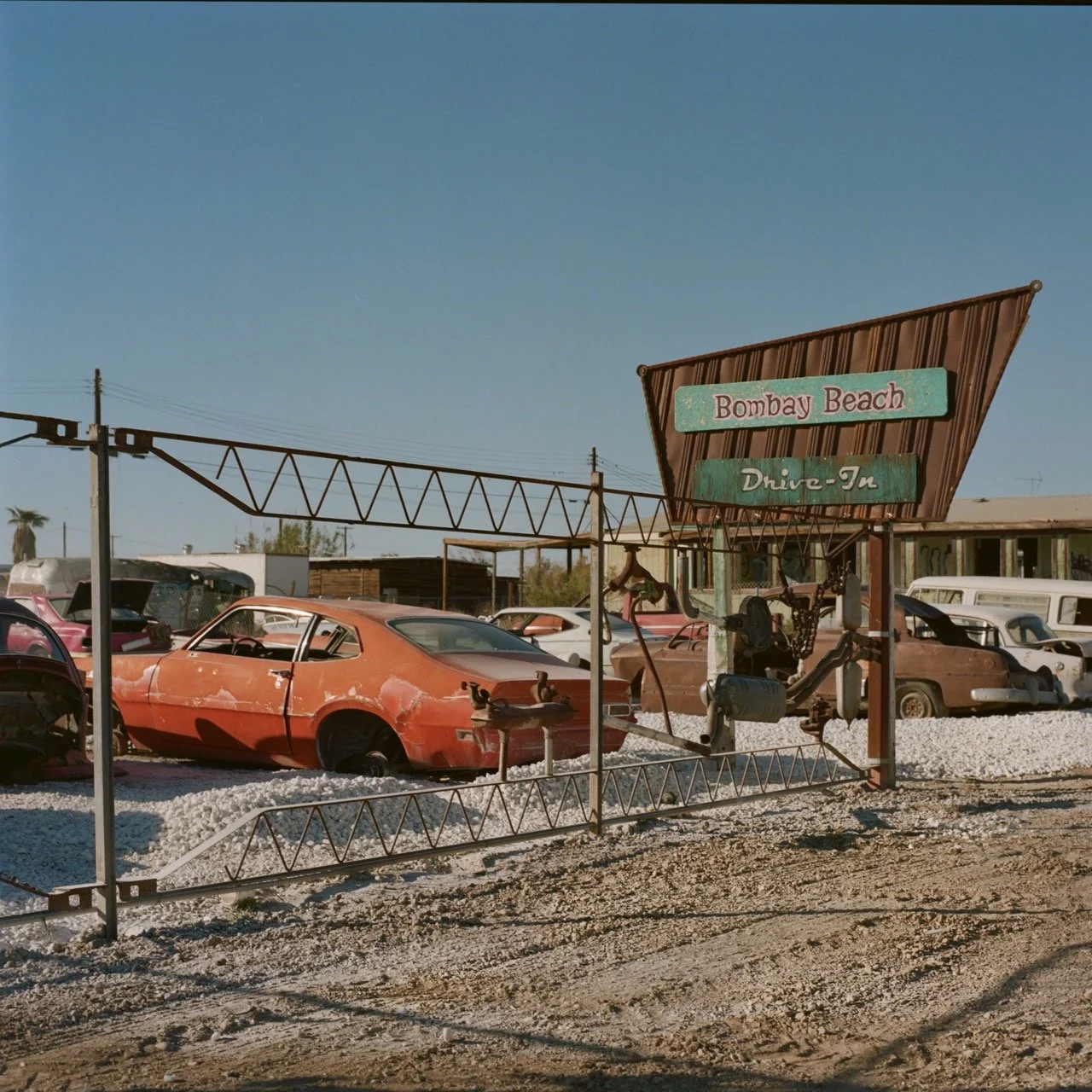 Old, abandoned cars are parked at Bombay Beach Drive-In, a vintage outdoor movie theater in a desert area, with a clear blue sky and a sign indicating the location.