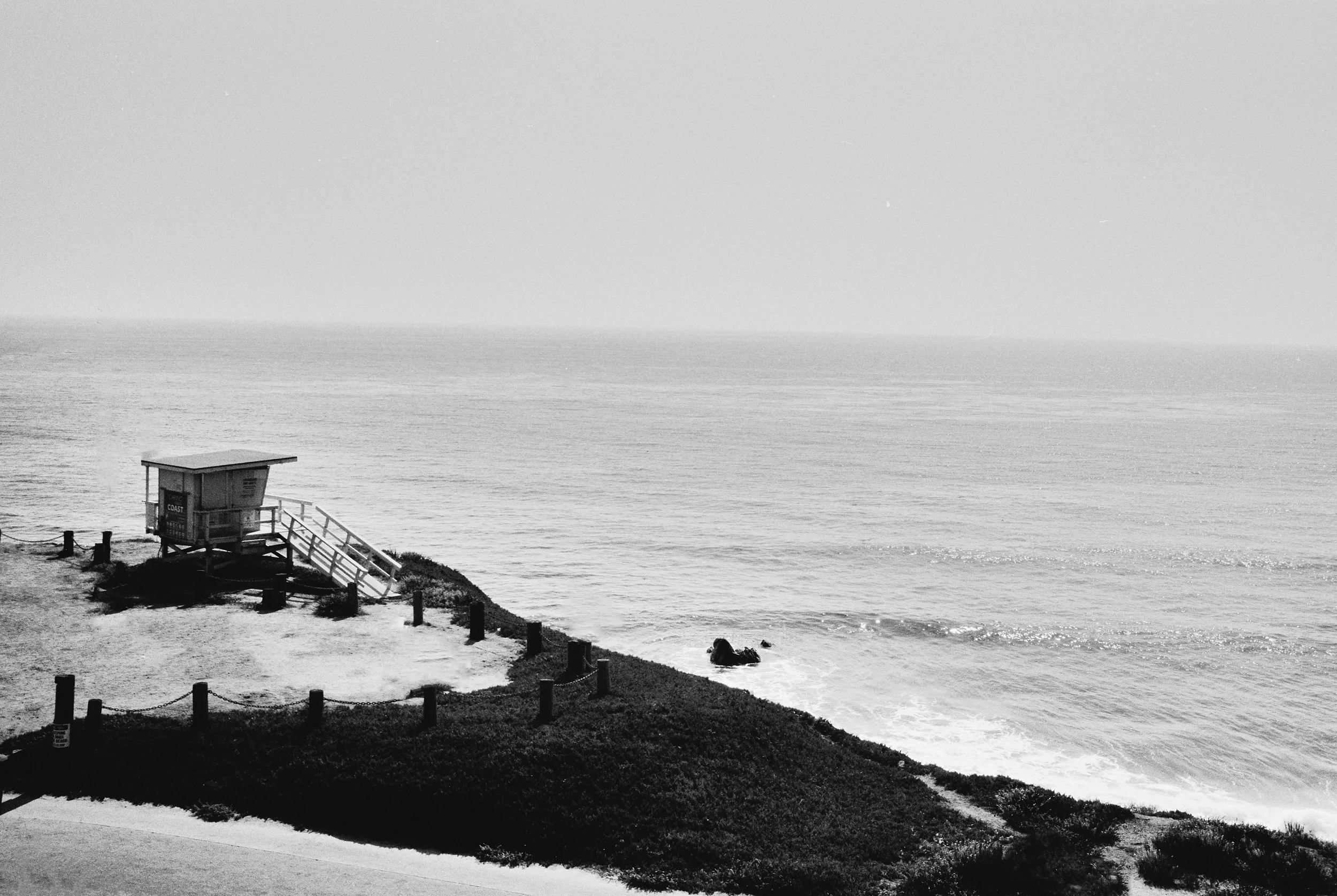 A black and white photo of a coastline with a lifeguard station on a small hill, surrounded by a fence and plants, overlooking the ocean.