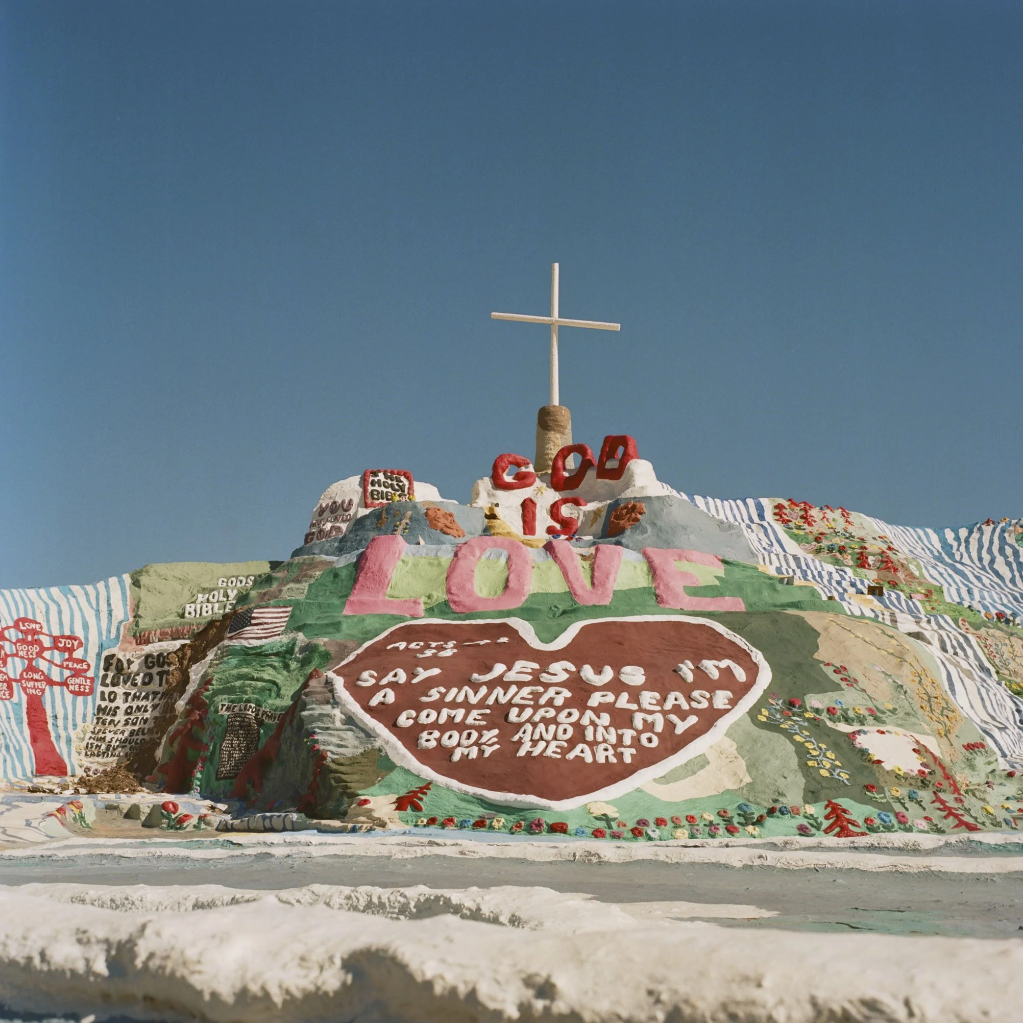 Colorful, layered sculpture with words, phrases, and symbols, including Christian cross at the top, and the word 'LOVE' in large letters. The sculpture features various decorations such as hearts, flags, and religious symbols, with the sky in the bac