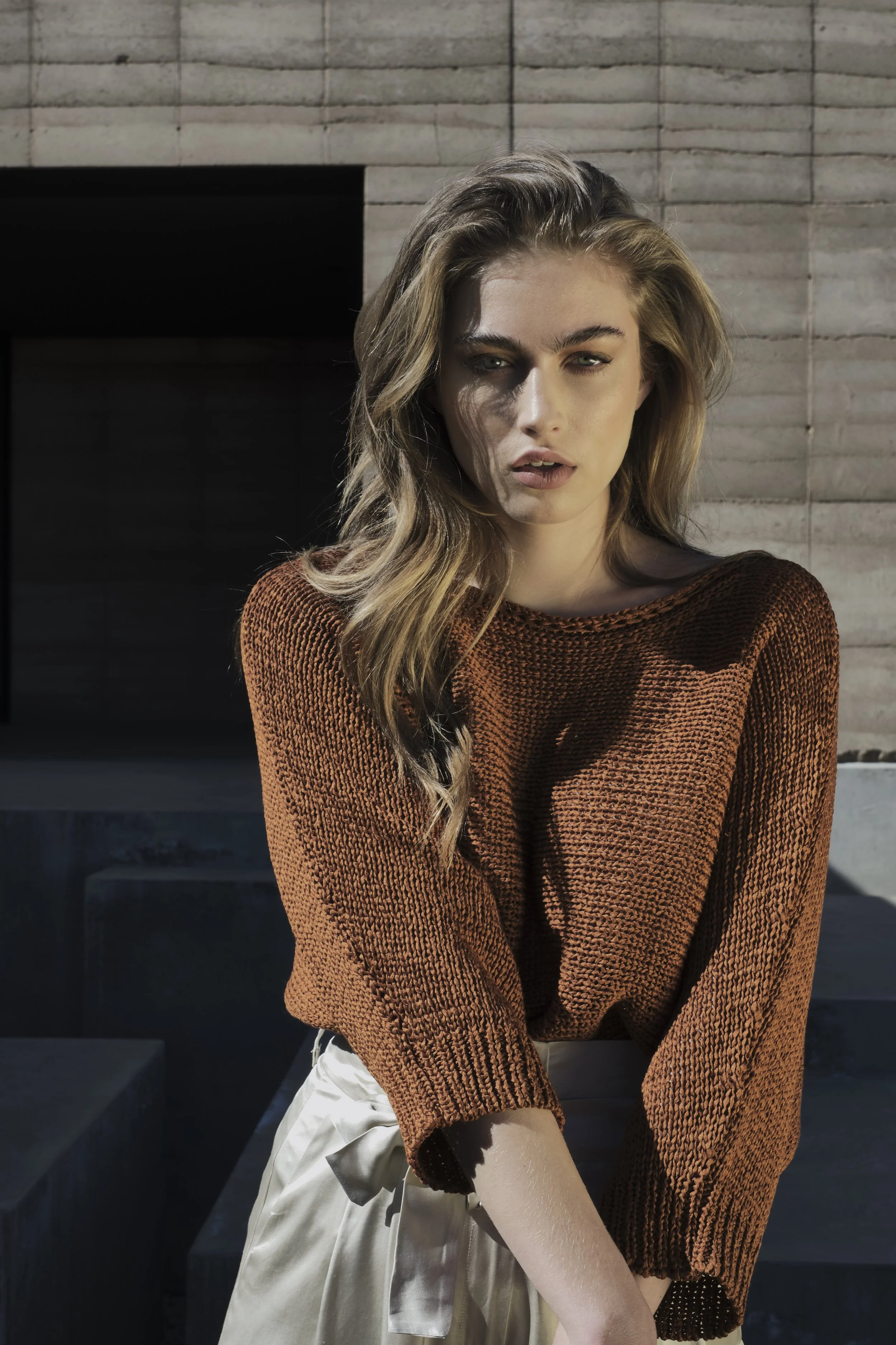 Portrait of a young woman with wavy blonde hair, wearing a brown knitted sweater and light-colored shorts, posing outdoors in front of a modern concrete building.