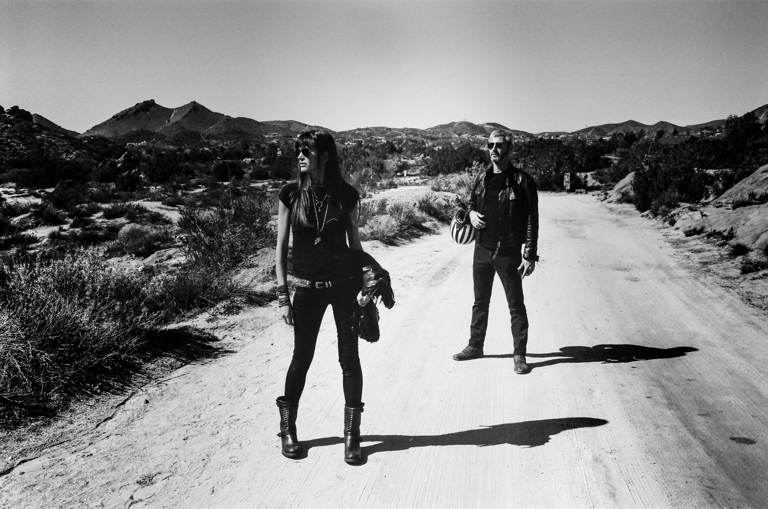 Two individuals walking on a dirt path in an arid, mountainous landscape. The woman in the foreground is wearing sunglasses, a black shirt, and boots, while the man in the background is wearing sunglasses, a leather jacket, and holding a helmet.