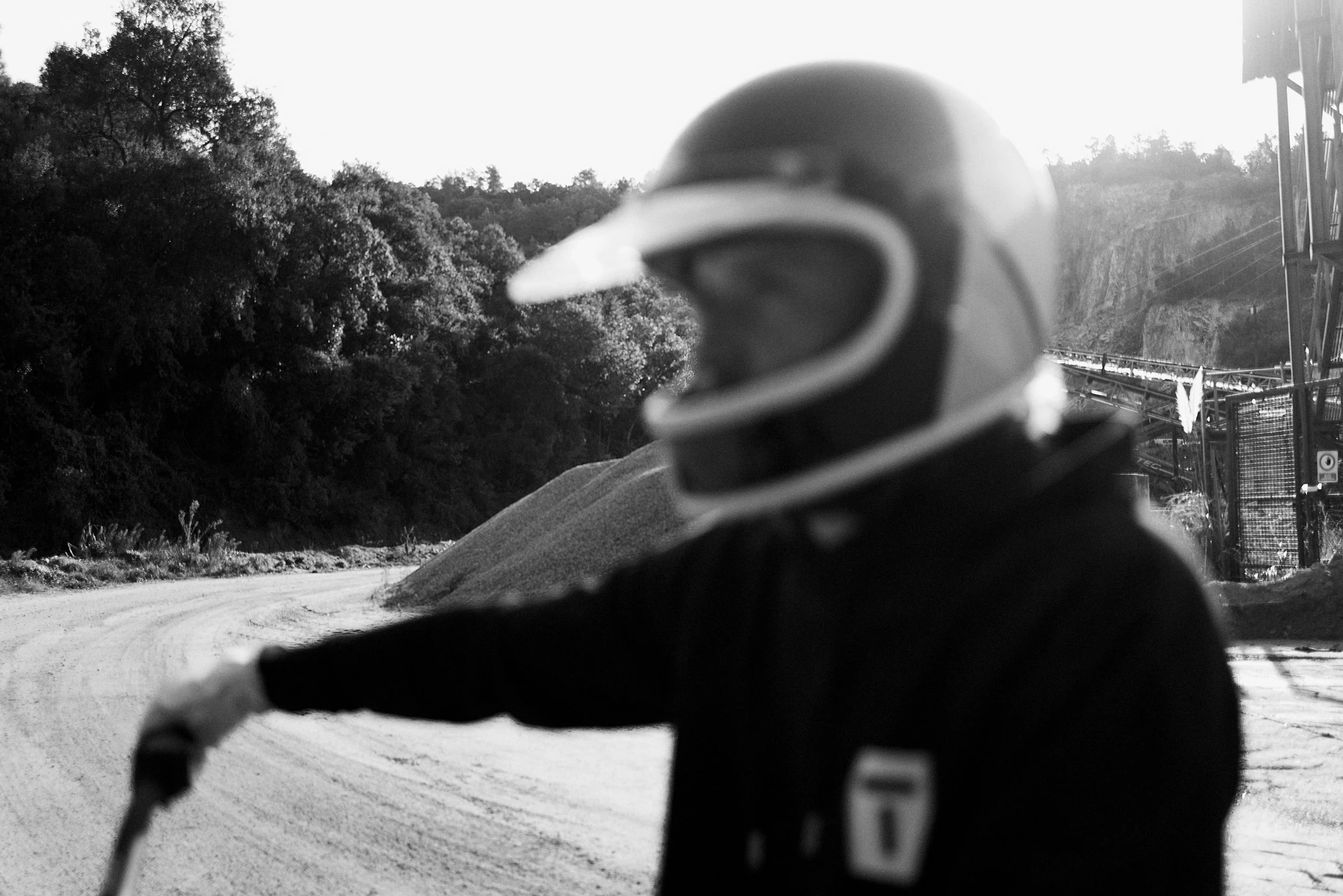 The image is a black and white photo of a person wearing a helmet, taken from behind. The person is outdoors, with trees and a hillside in the background, and a dirt road in front of them.