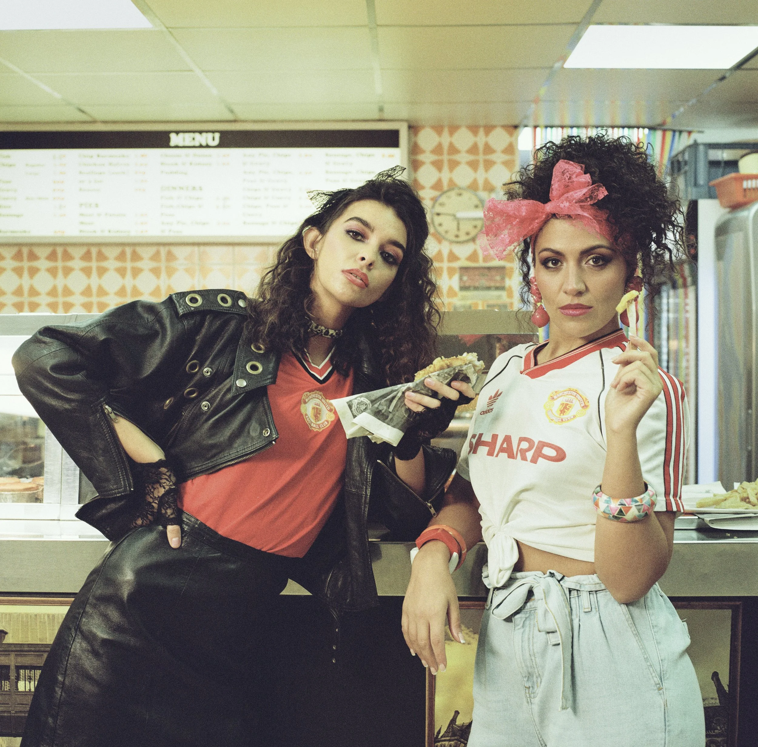 Two women pose inside a fast-food restaurant. The woman on the left has dark wavy hair, a leather jacket, and makes a peace sign. The woman on the right has curly hair with a pink headscarf, and wears a white sports jersey with red and yellow accents