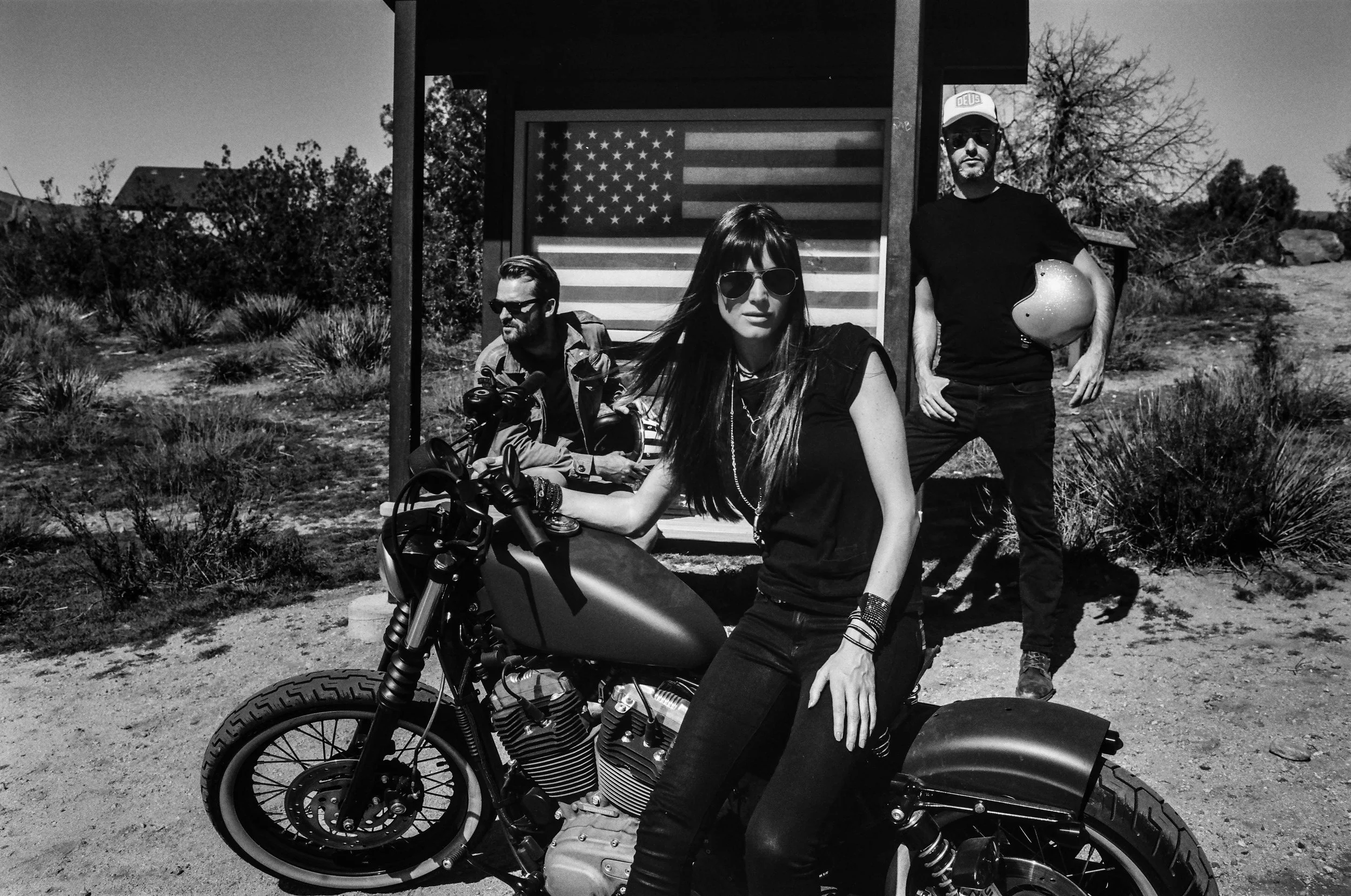 Three people posing outdoors with a motorcycle and an American flag in the background. Two men and one woman, dressed in casual clothing and wearing sunglasses, in a natural setting with trees and bushes.