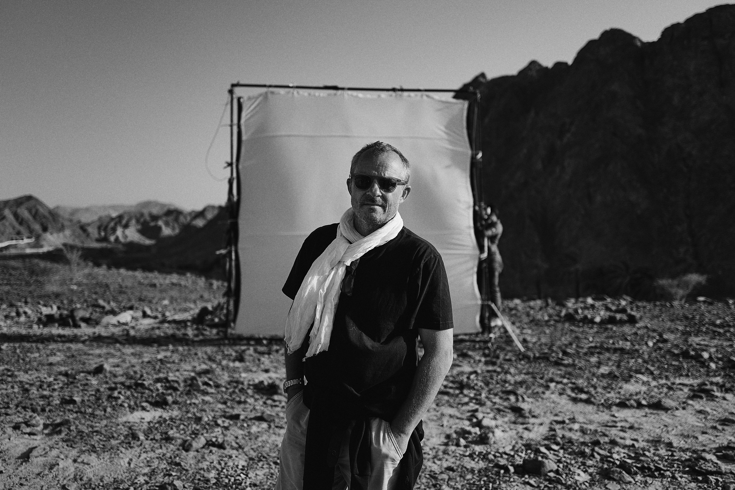 A man with sunglasses and a scarf standing outdoors in a rocky desert landscape with mountains in the background.