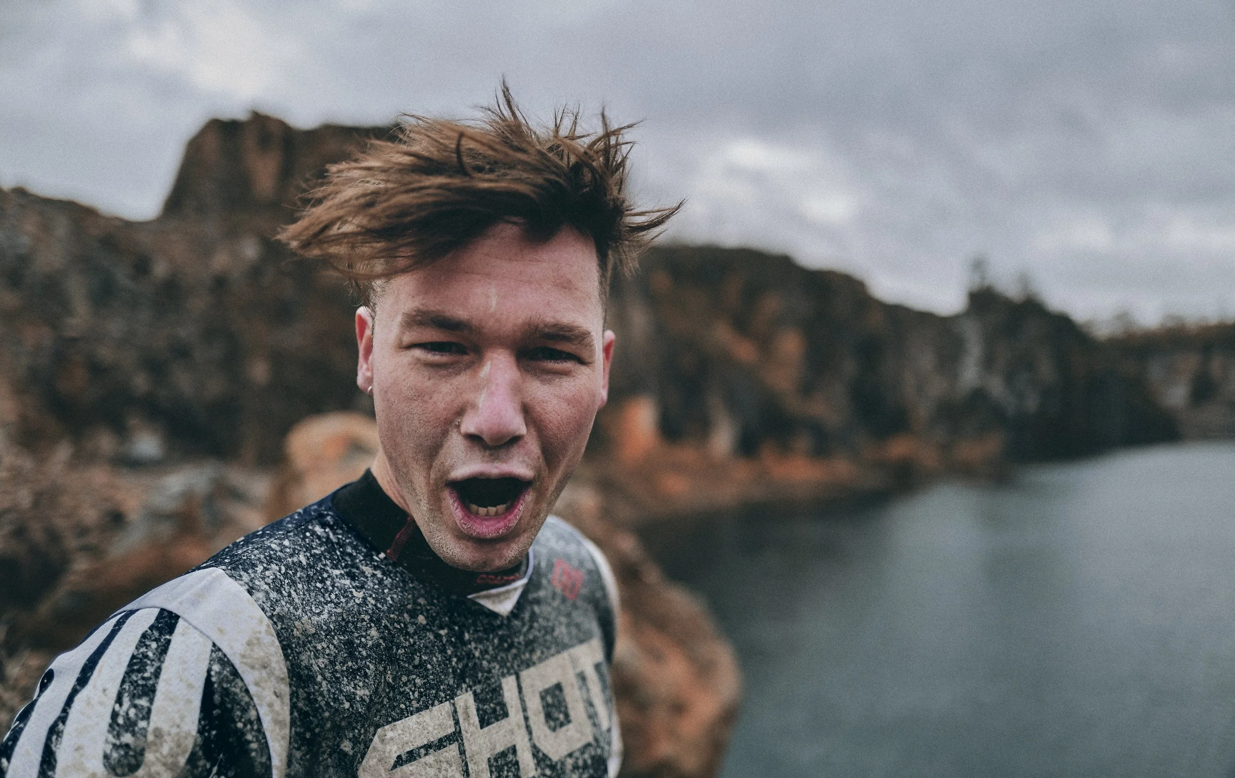 Young man with short brown hair, screaming or shouting, wearing a black and white motocross jersey, outdoors near rocky cliffs and water under cloudy sky.
