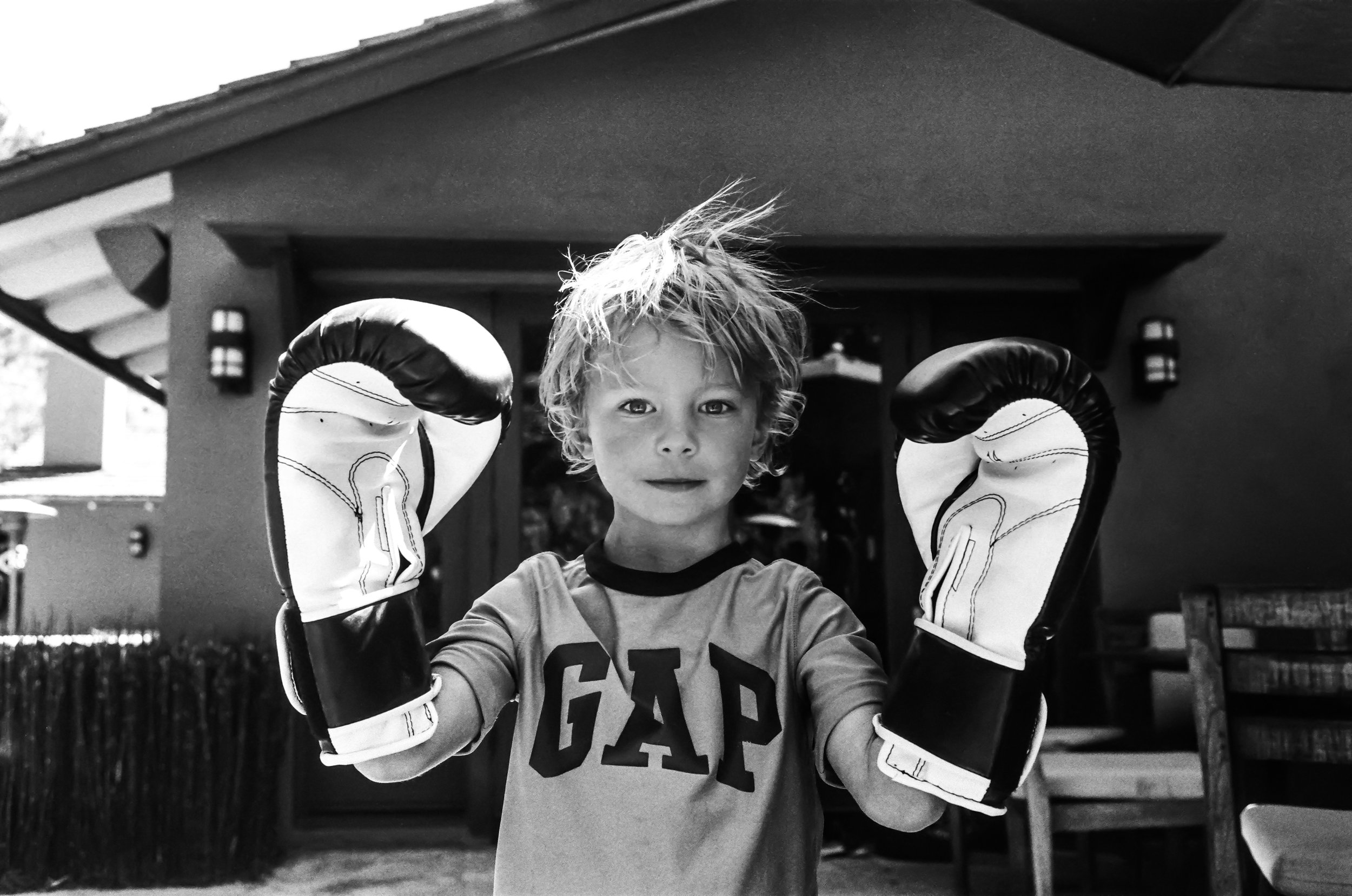 A young boy with messy hair wearing boxing gloves, standing outdoors in front of a house, looking into the camera.