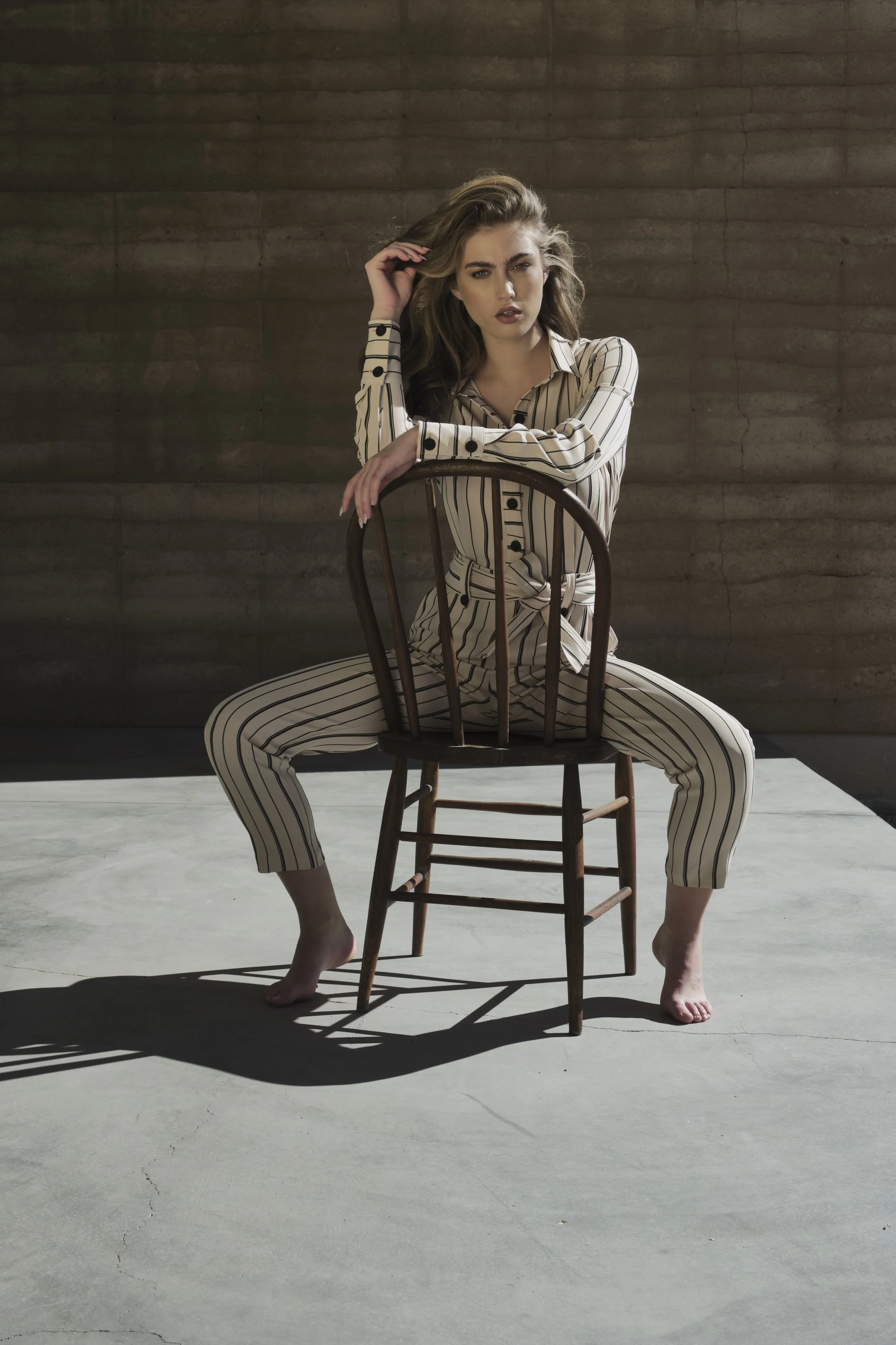 A woman sitting on the floor behind a wooden chair, wearing striped pajamas, with one hand on her head and the other resting on the chair. Her feet are on the ground, and her face shows a serious or contemplative expression. The background is a plain
