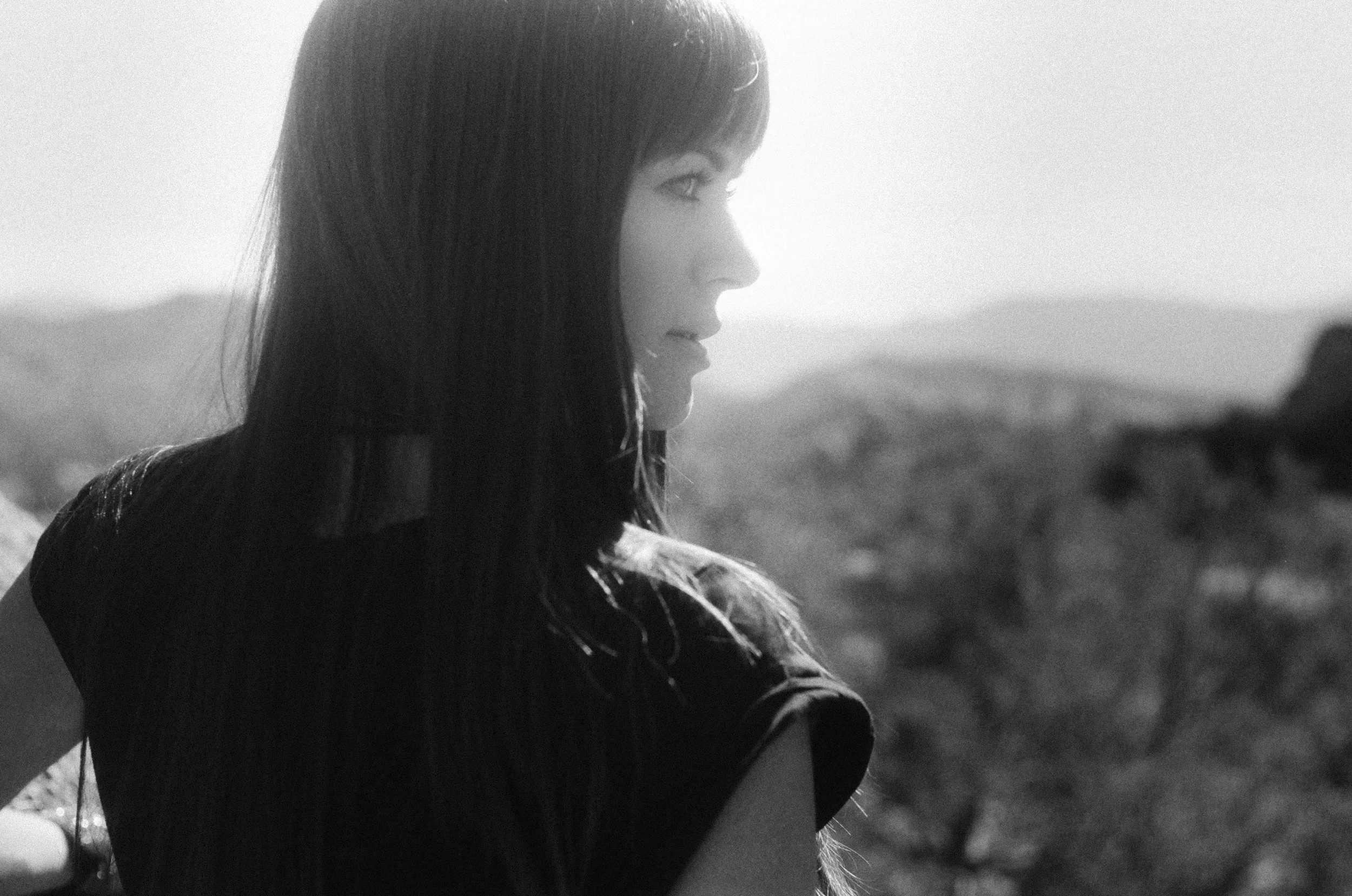 A black-and-white photo of a young woman with long dark hair, looking sideways and smiling softly, with a blurred natural landscape background.