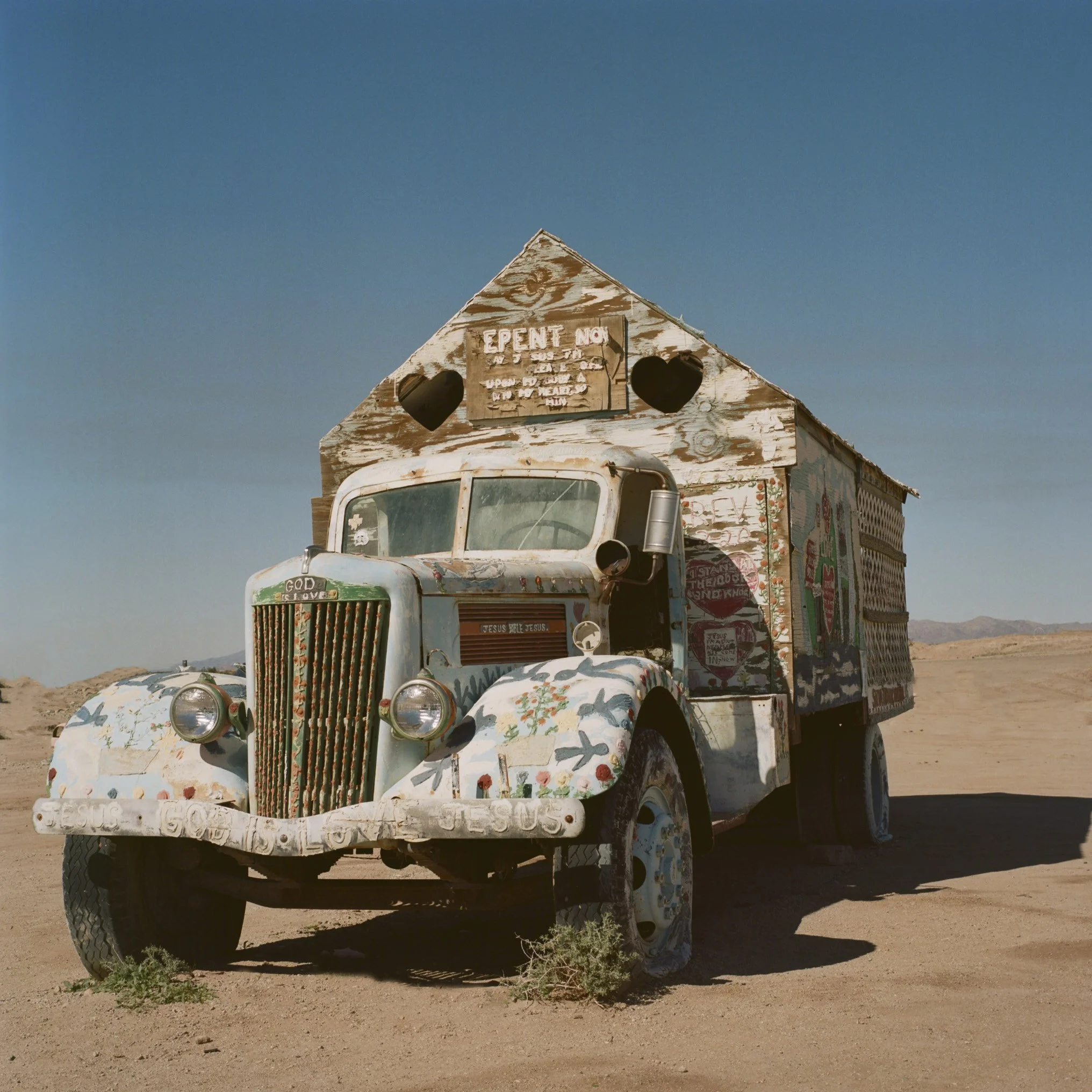An old, weathered truck painted with colorful flowers and religious messages, sitting in a desert landscape with mountains in the background.