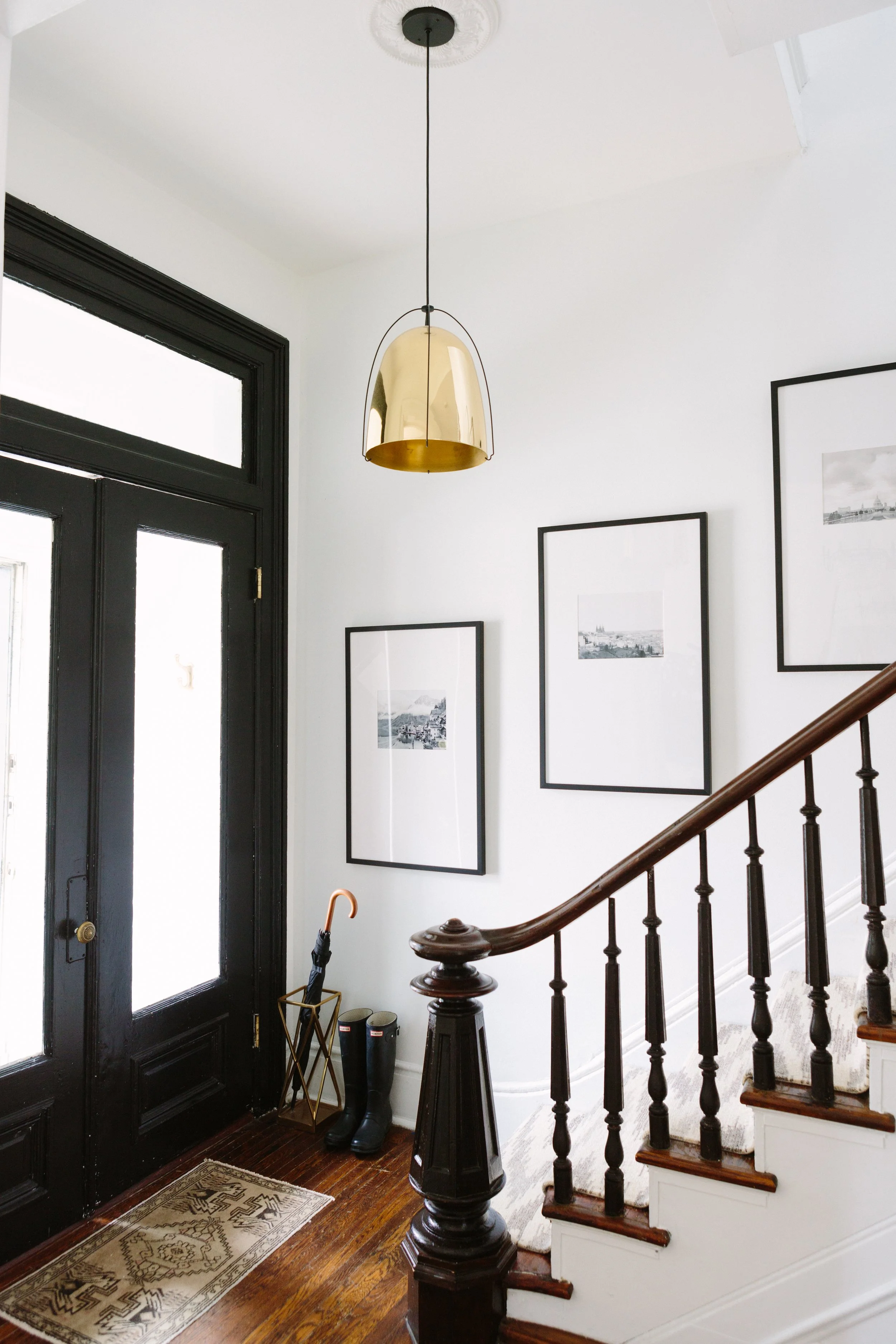 Staircase gallery wall with a vertical linear arrangement of three matching thin black frames ascending along a dark wood banister. White walls contrast with the dark frames and rich hardwood floors. A warm brass pendant light anchors the entryway, w