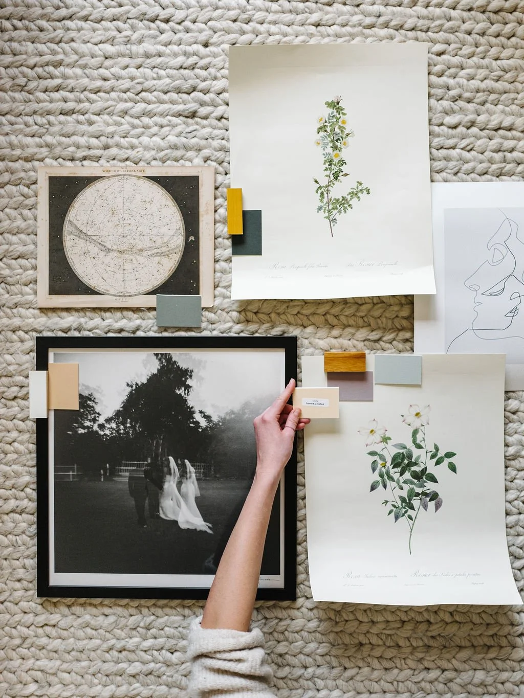 Art curation mood board with a hand holding a black framed landscape photo surrounded by botanical prints, fabric swatches, paint chips, and a vintage map on a woven surface
