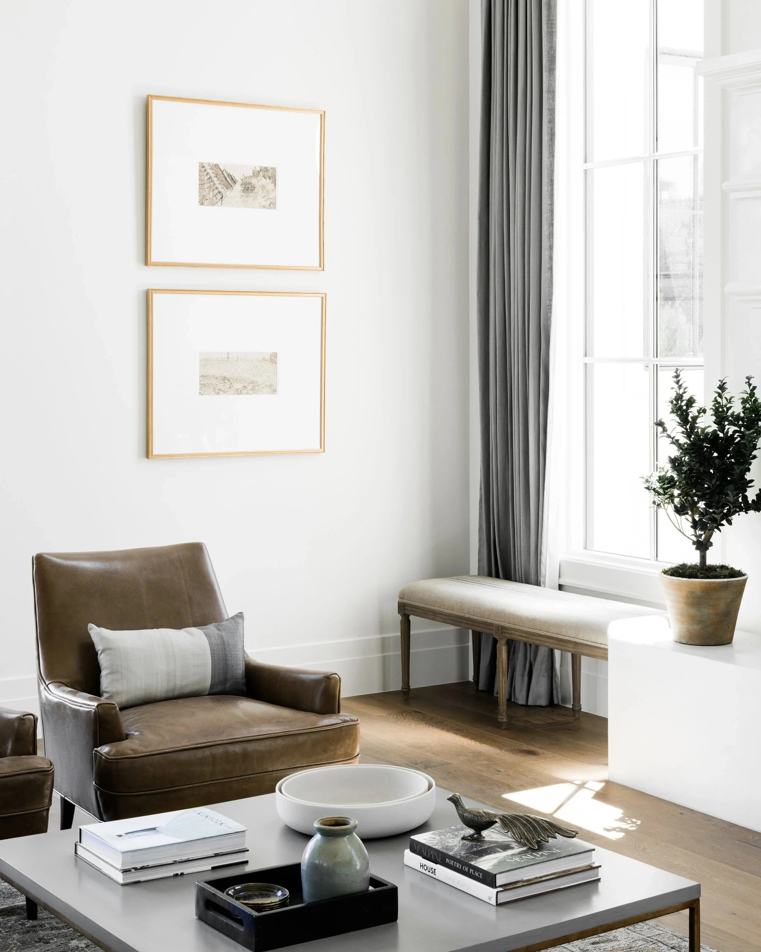 Minimalist living room with two stacked thin gold frames displaying landscape prints on a white wall above a neutral armchair and coffee table