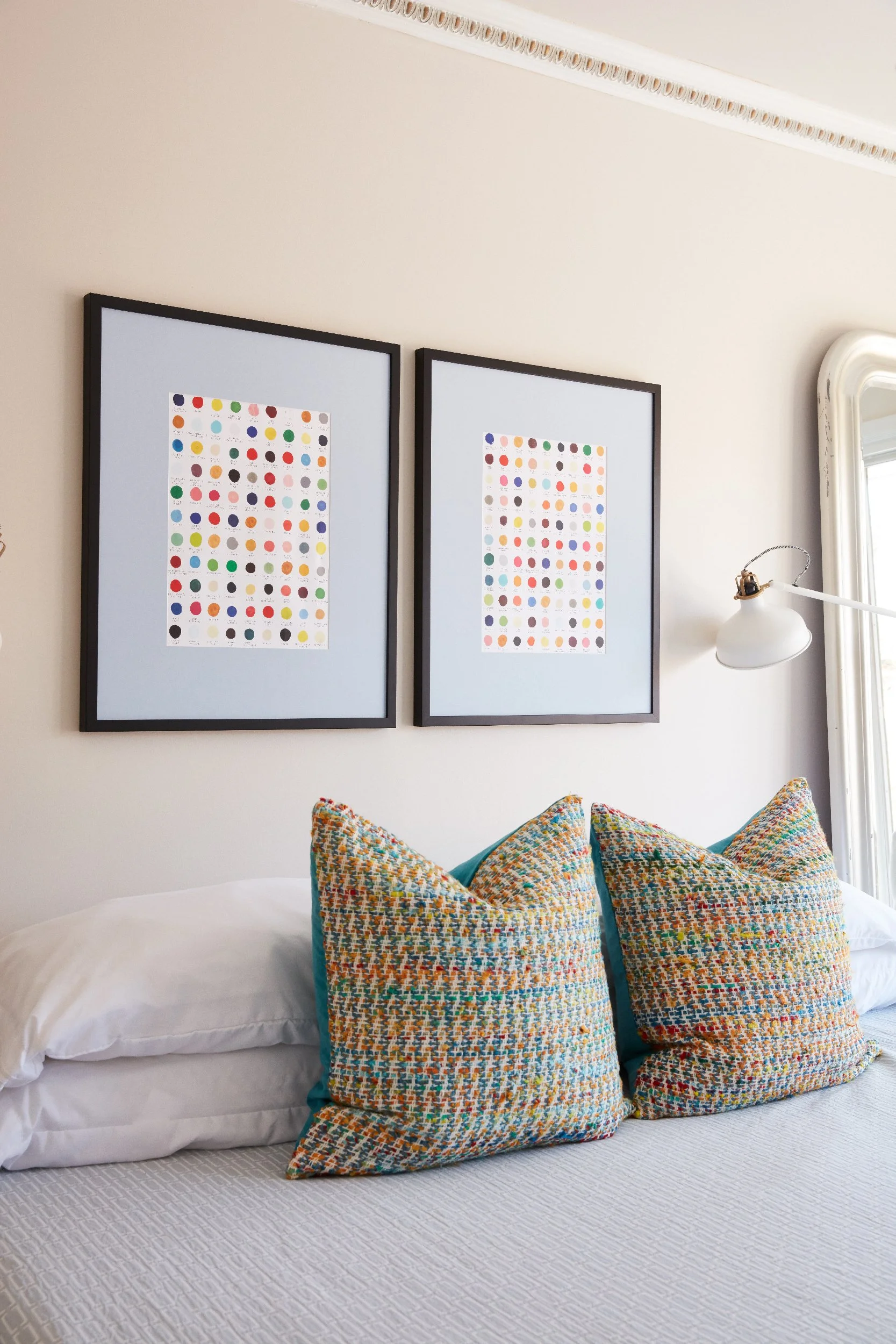 Two matching black frames with blue mats displaying colorful dot grid prints hung side by side above a bed with multicolor woven pillows