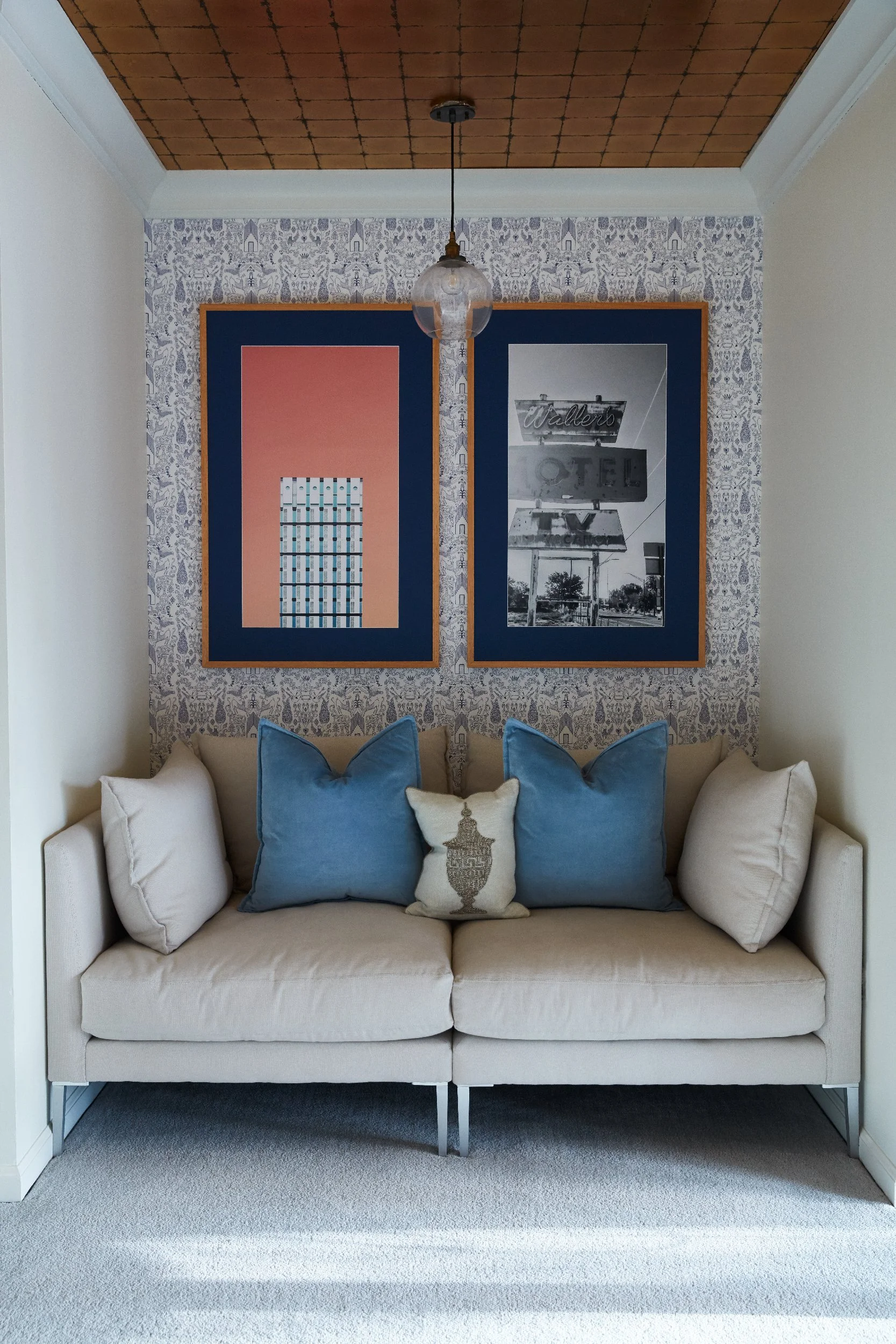 Residential living room with two large navy blue frames displaying a coral abstract print and black and white architectural photography above a neutral sofa with blue accent pillows, set against bold patterned wallpaper