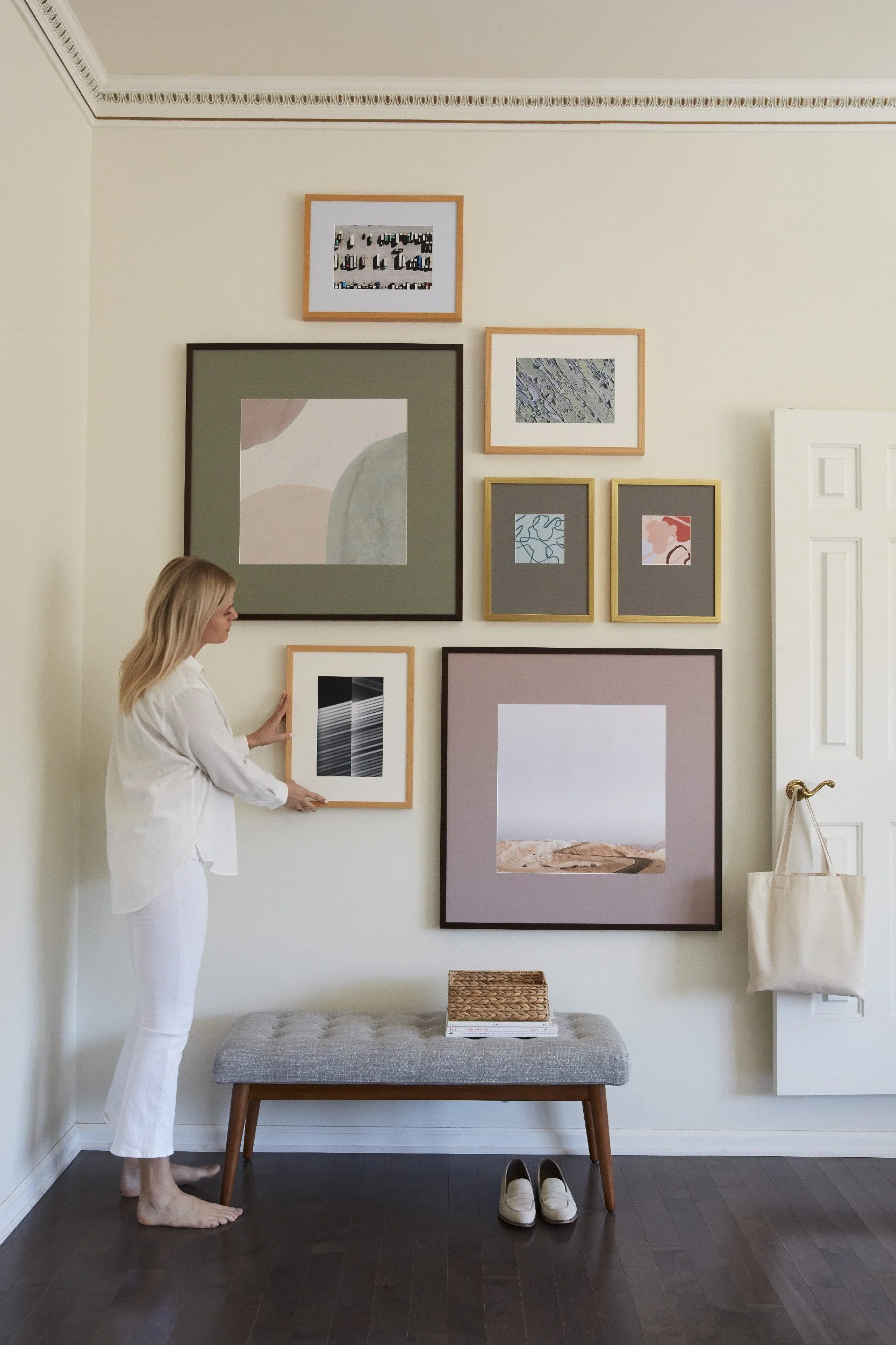 Woman arranging a gallery wall with mixed gold and dark frames featuring abstract and nature-inspired art prints above a tufted bench