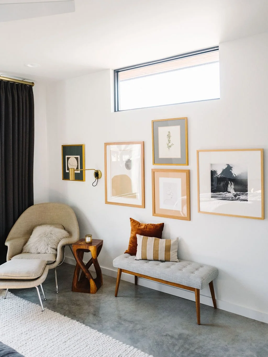 Curated gallery wall in a bright living room featuring an asymmetrical arrangement of five framed art pieces in warm gold and natural wood frames. Artwork includes abstract prints, a landscape, and a dark botanical piece, displayed above a neutral up