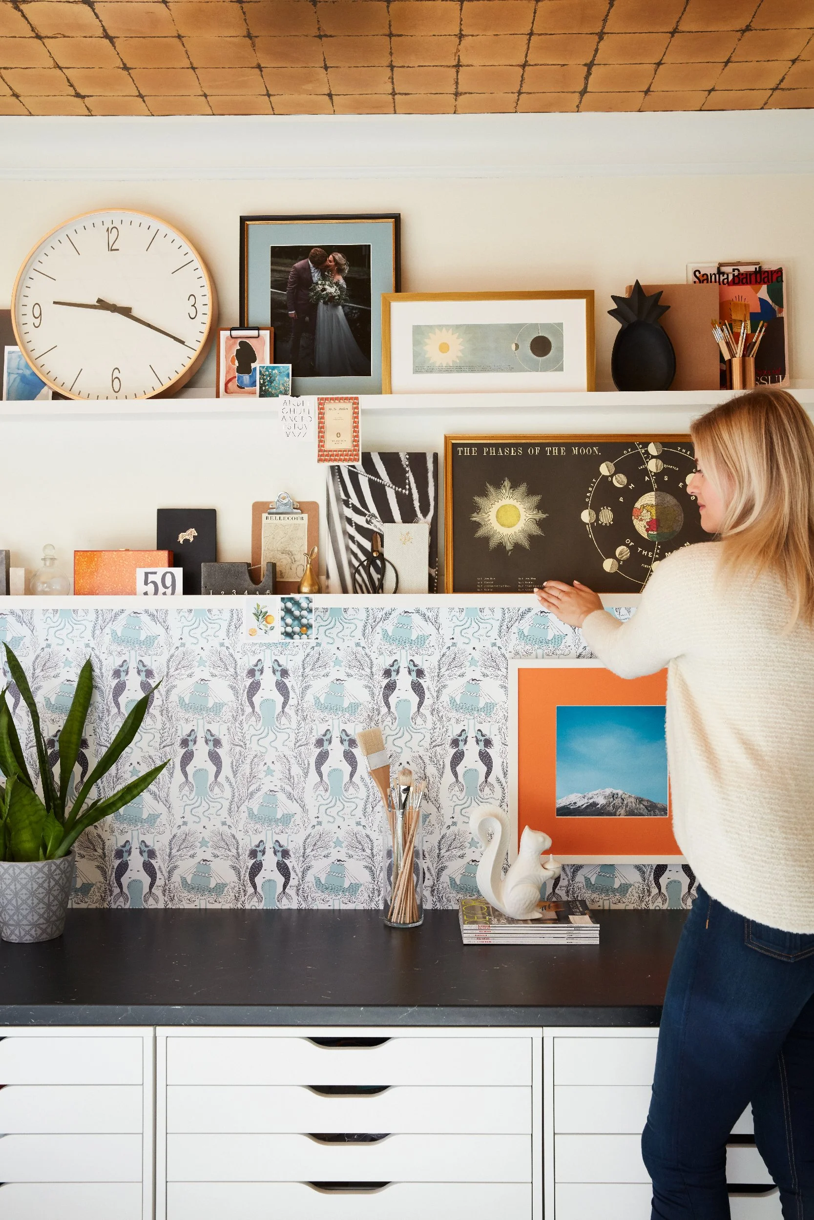 Woman curating an eclectic gallery wall and shelf display featuring vintage astronomy prints, a wedding photo, framed artwork, a large wall clock, and layered decorative objects on patterned wallpaper