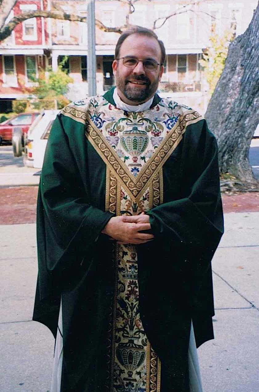 A man in a religious or ceremonial robe standing outdoors on a sidewalk, with trees and residential buildings in the background.