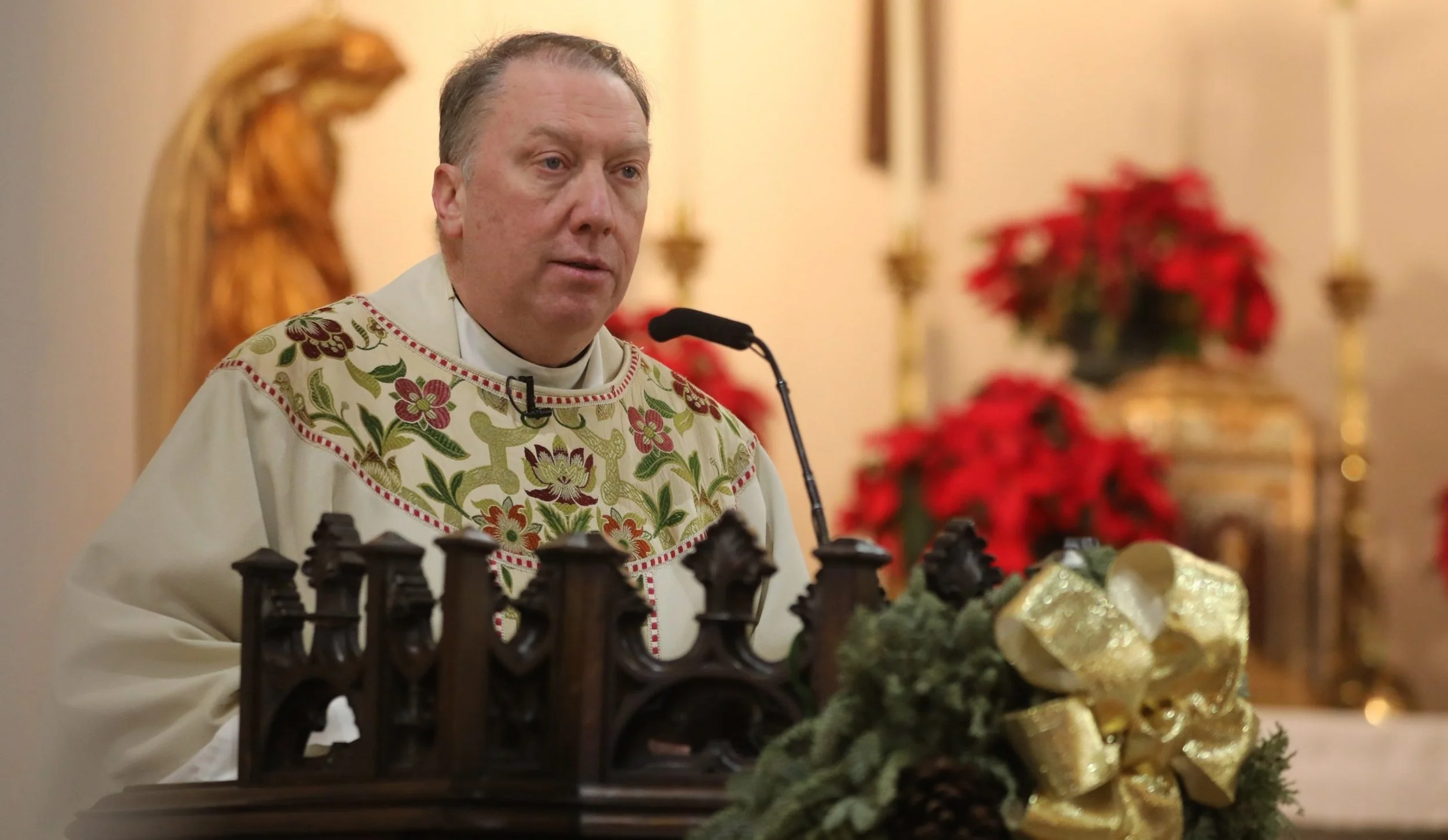 A priest standing at a lectern, dressed in religious vestments with floral embroidery, speaking into a microphone during a Christmas service, with poinsettias, a poinsettia-shaped decoration, and a golden angel in the background.
