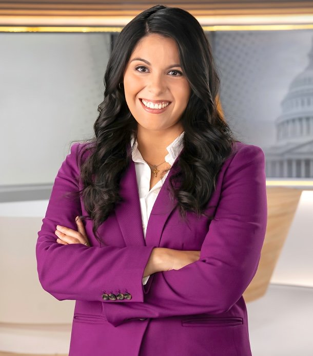A smiling woman with long dark hair wearing a purple blazer with her arms crossed, standing in a professional studio setting with a blurred background of the U.S. Capitol.