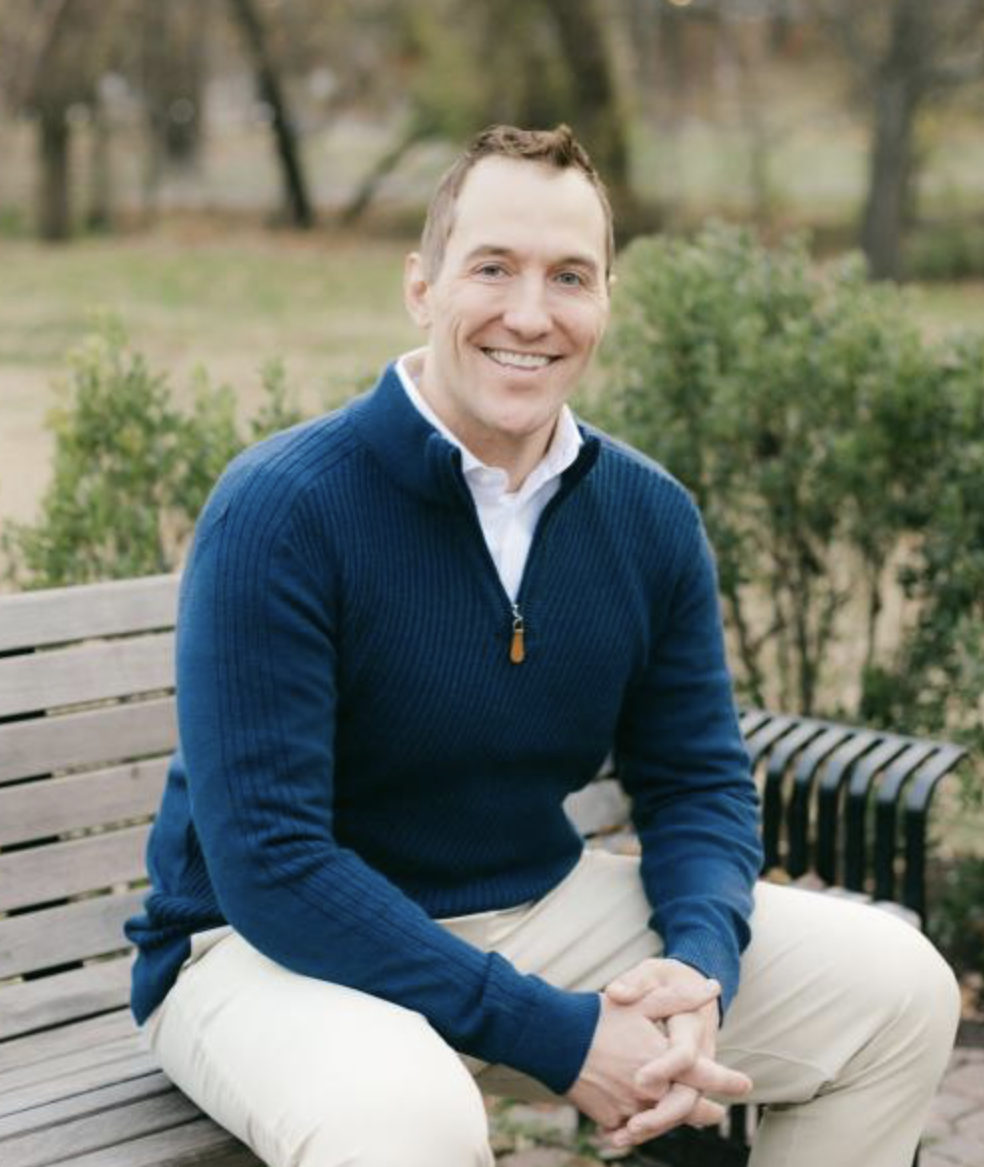 A smiling man sitting on a park bench with greenery and trees in the background, wearing a blue sweater and beige pants.
