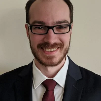 A young man with glasses, a beard, and short hair, wearing a suit with a white shirt and a maroon tie, smiling at the camera.