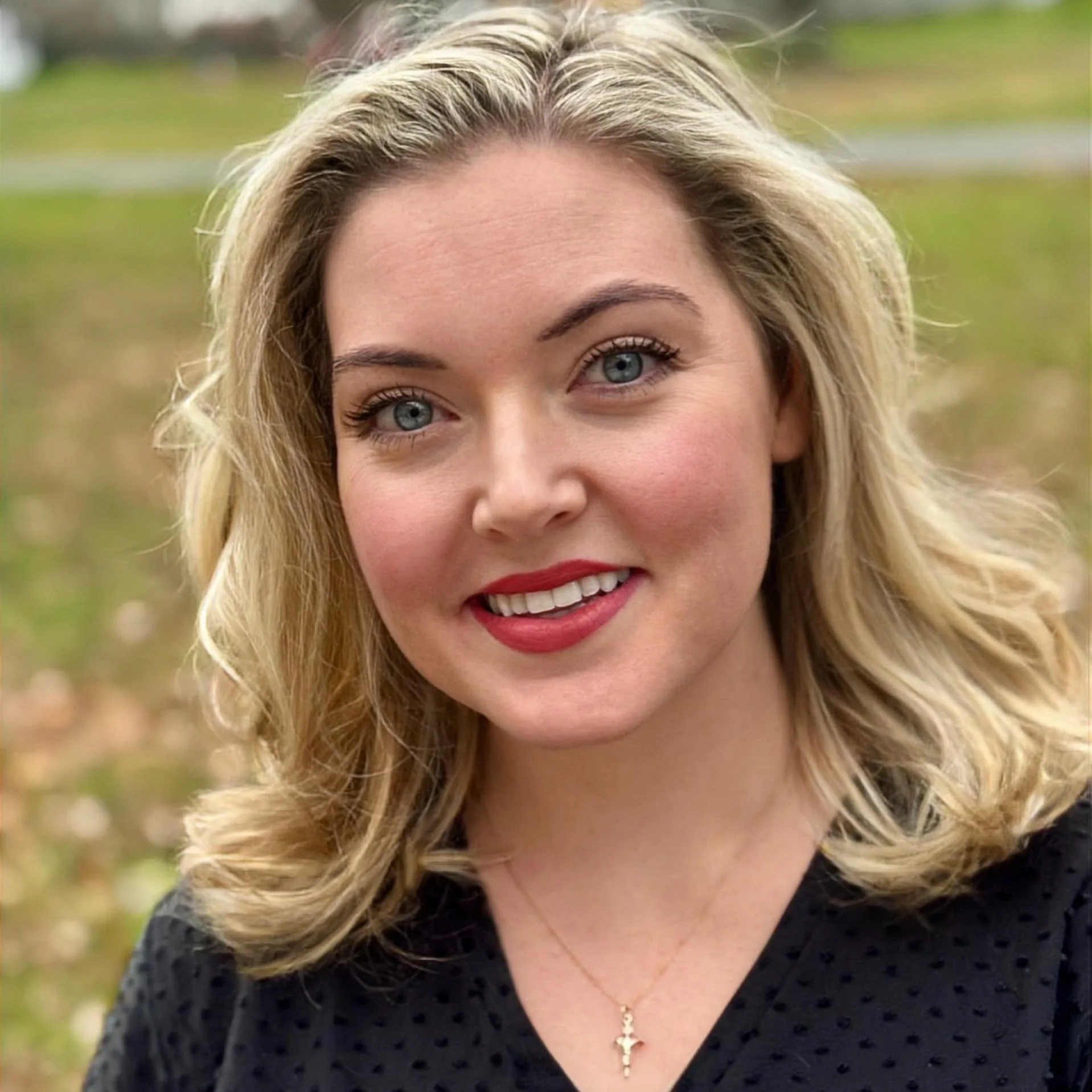 Close-up of a smiling blonde woman with blue eyes, wearing red lipstick, a black top, and a gold cross necklace, outdoors with a blurred grassy background.