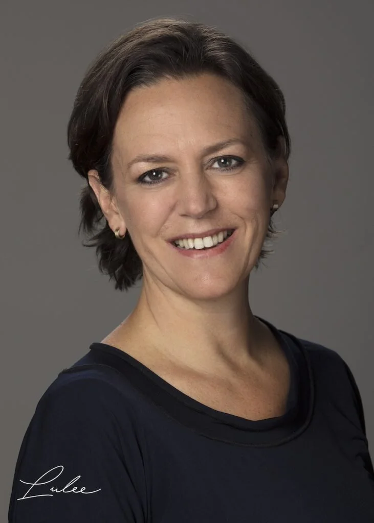 A woman with short brown hair, wearing a black top, smiling at the camera, against a gray background.