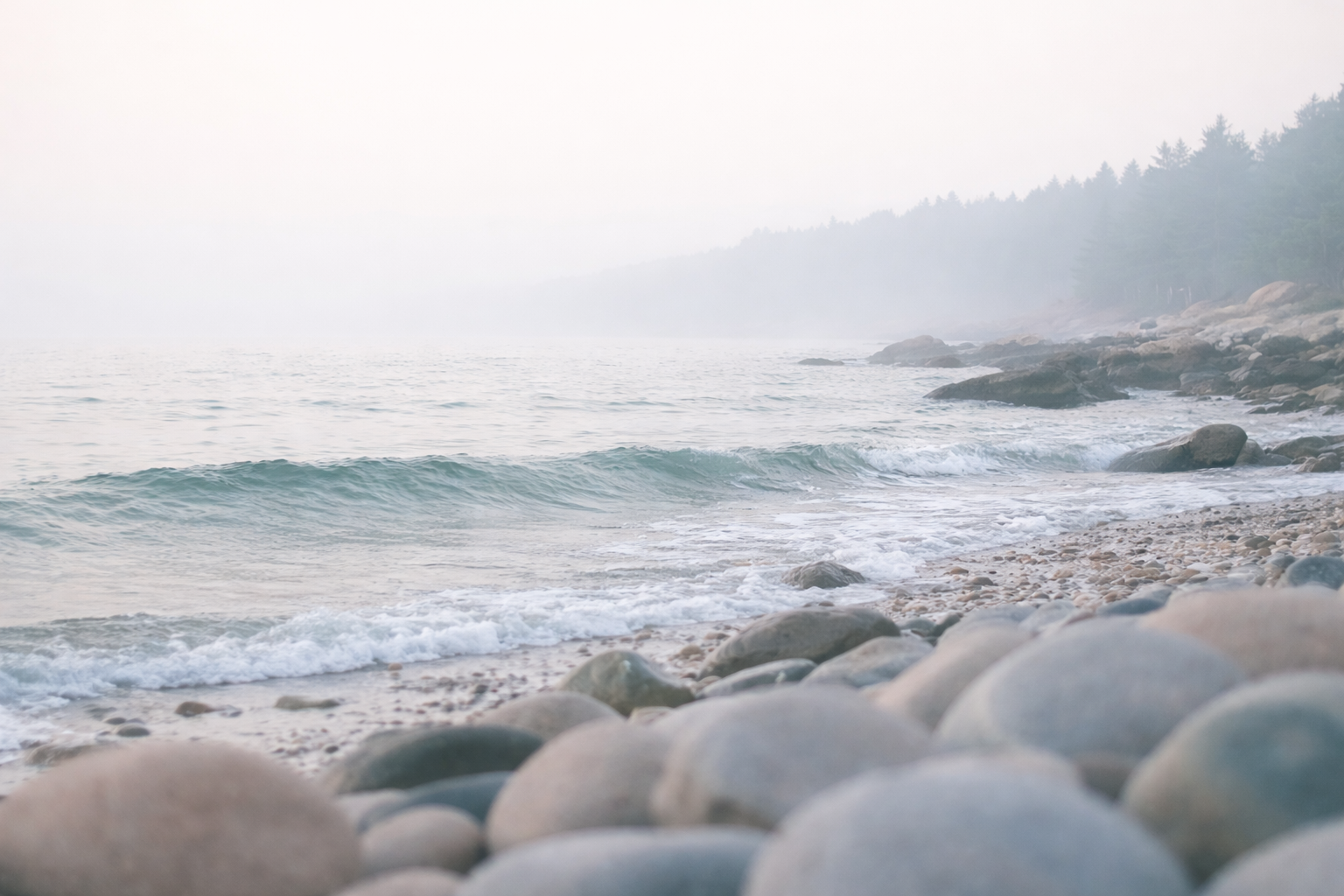 A rocky beach with small waves and a misty forested coastline in the distance.