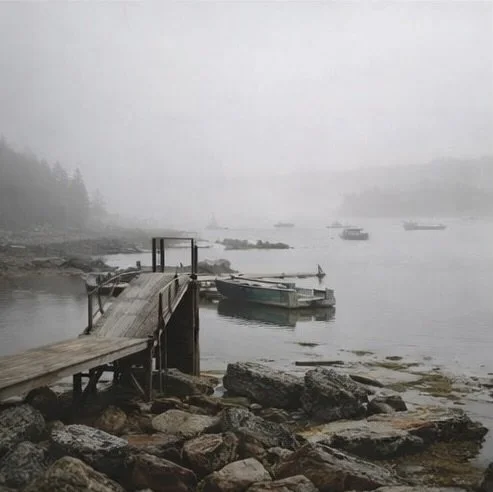 A foggy ocean cove scene with a wooden dock extending over rocks, with boats tied to the dock and floating in the water.