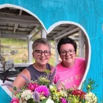Two women smiling behind a large bouquet of colorful flowers, in a bright indoor setting.