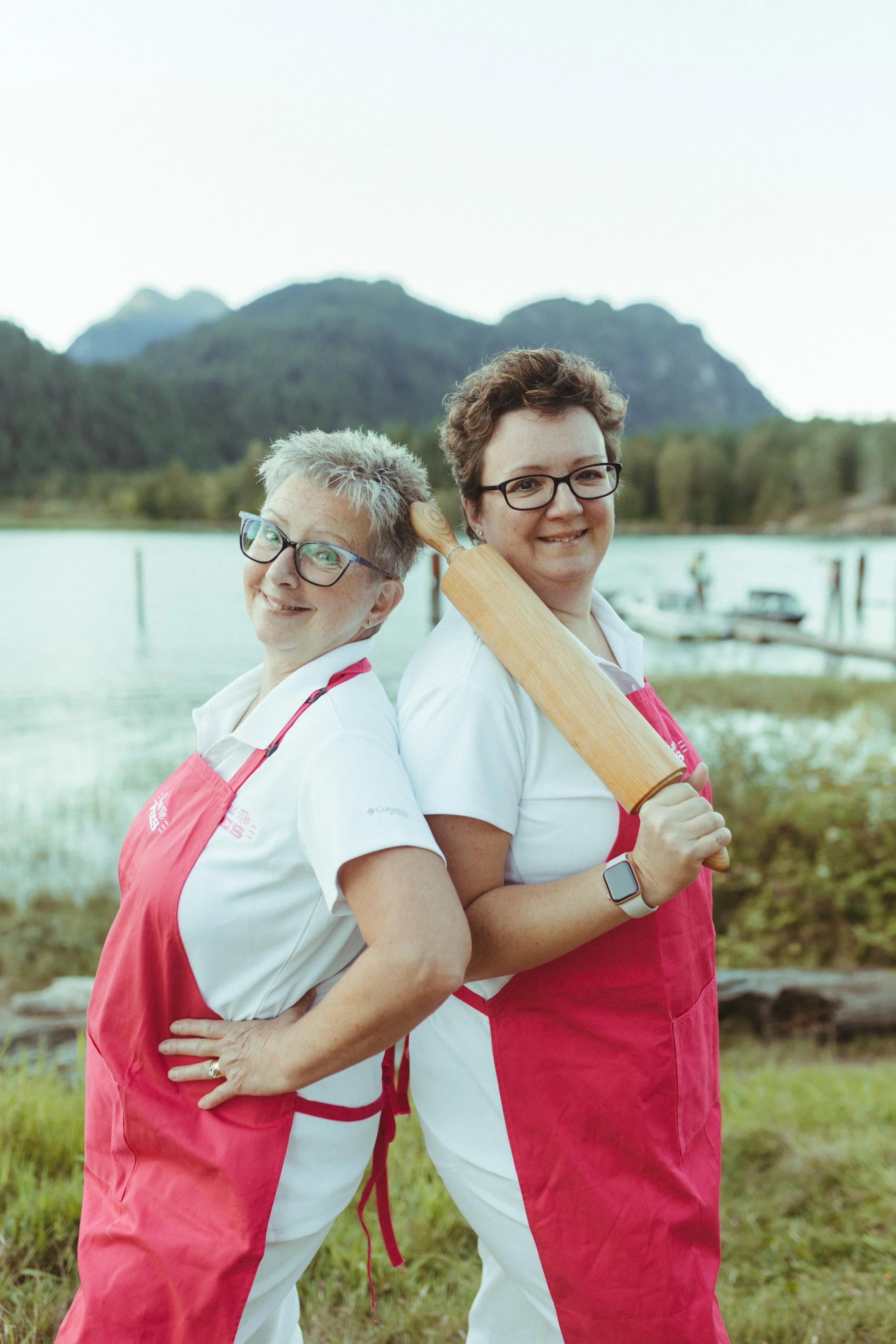 Two women in white shirts and red aprons standing back to back outdoors with a lake and mountains in the background, one woman holding a rolling pin.